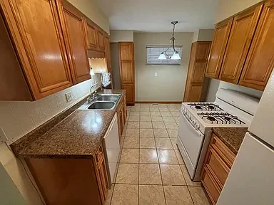 Empty kitchen with wooden cabinets, granite countertops, white appliances, tiled floor, and a small window with blinds.