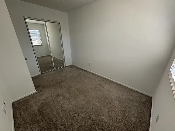 Empty bedroom with beige carpet, white walls, a window with blinds, and a mirrored closet door.