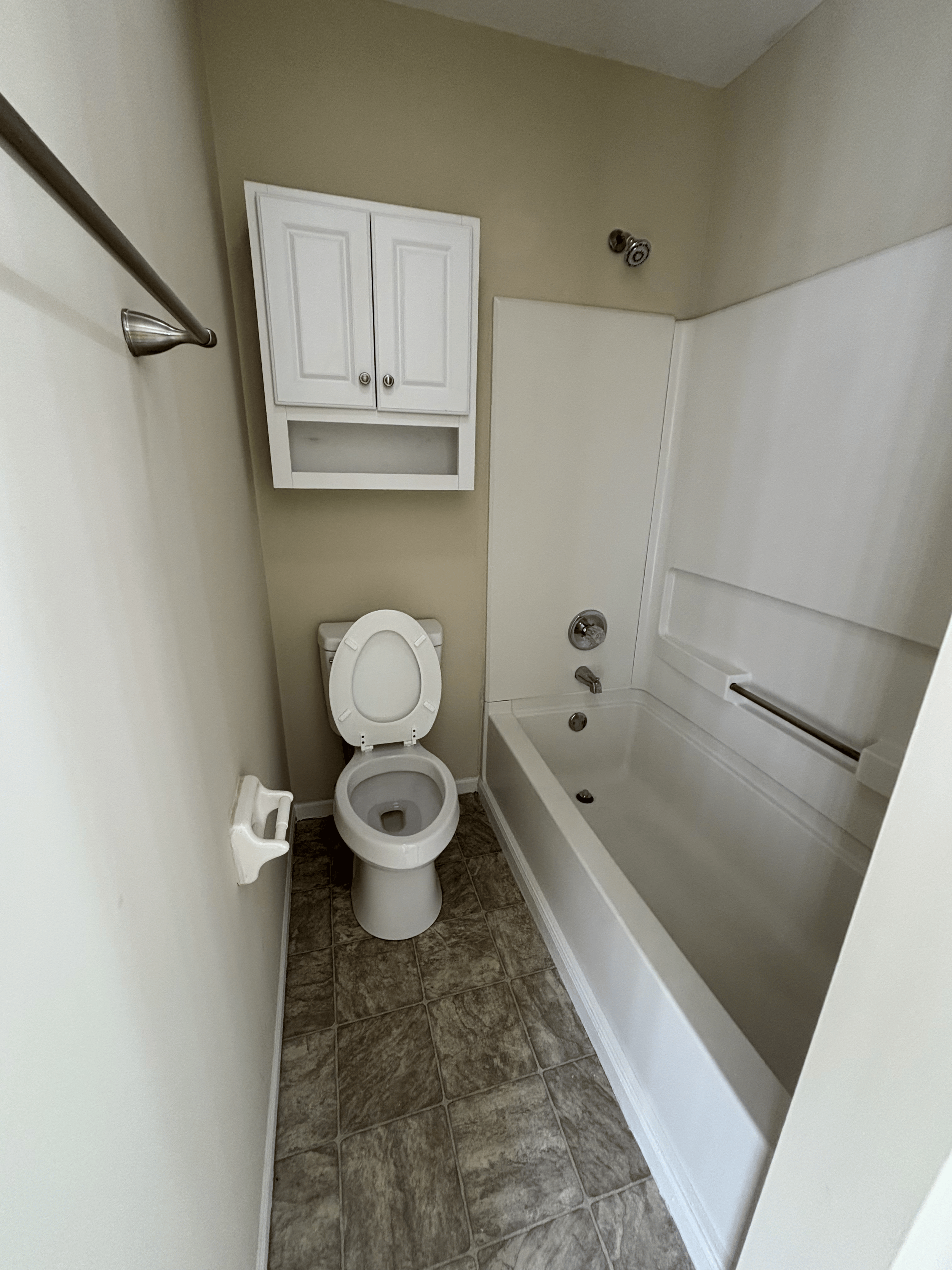 Small bathroom with a toilet, a bathtub with a grab bar, a white cabinet above the toilet, and tile flooring.