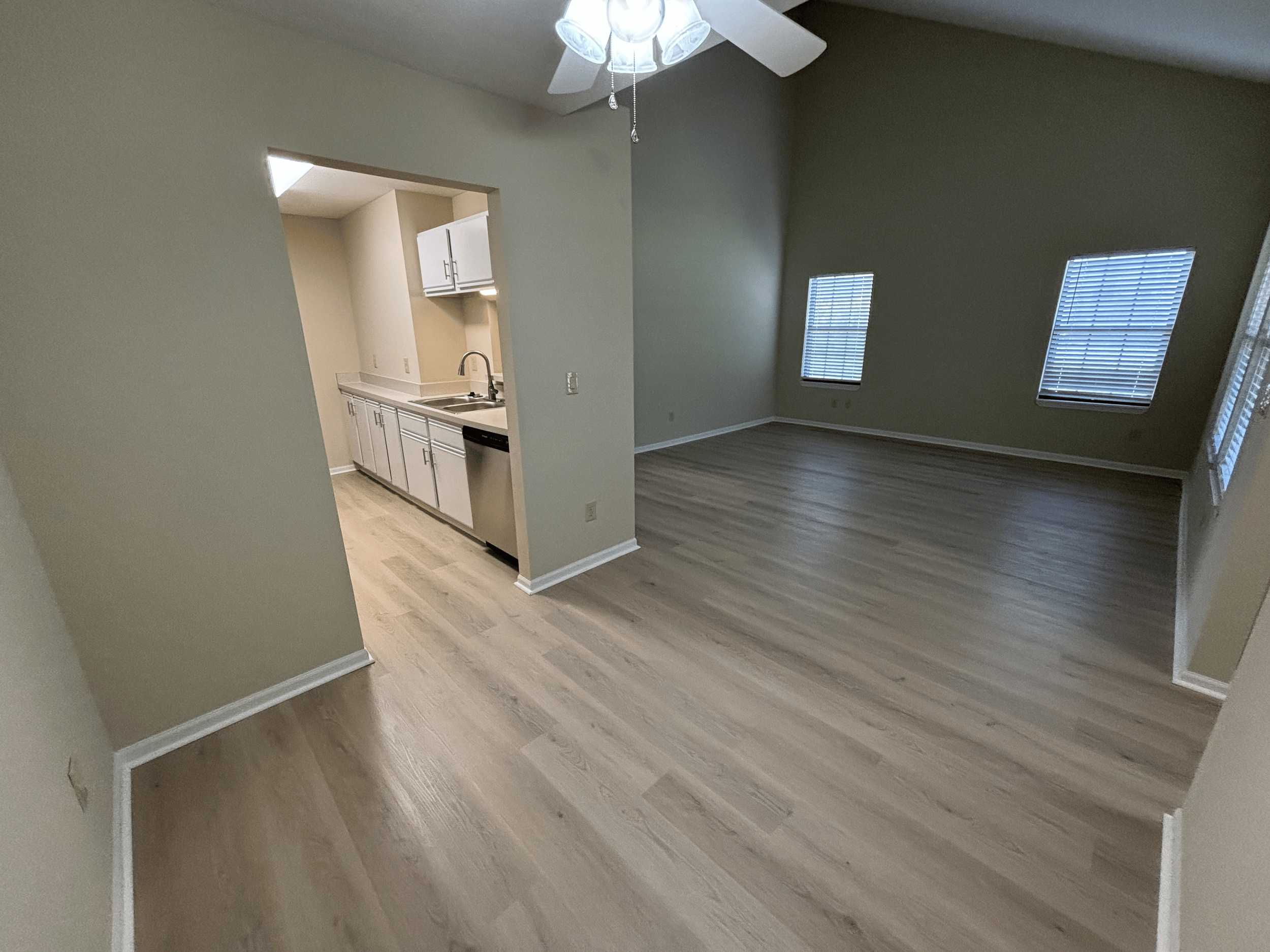 Empty living room with light-colored wooden floors and three windows with blinds, adjacent to a kitchen with white cabinets, a sink, and a ceiling fan.