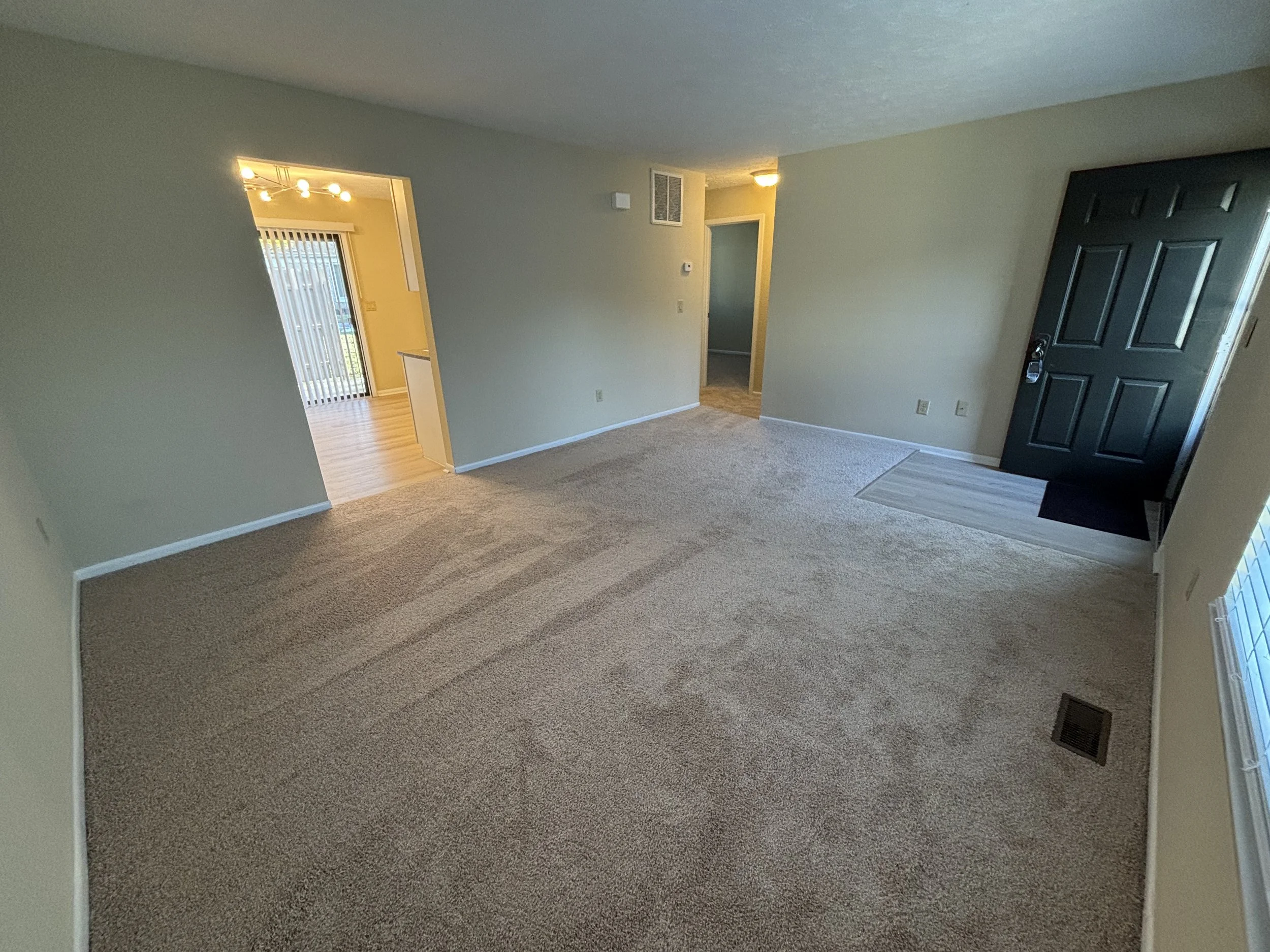 Empty living room with beige carpet, light-colored walls, and a black front door. A brick-patterned vinyl mat is in front of the door, and a vent is on the floor near the window. An open doorway leads to another room with sliding glass doors.