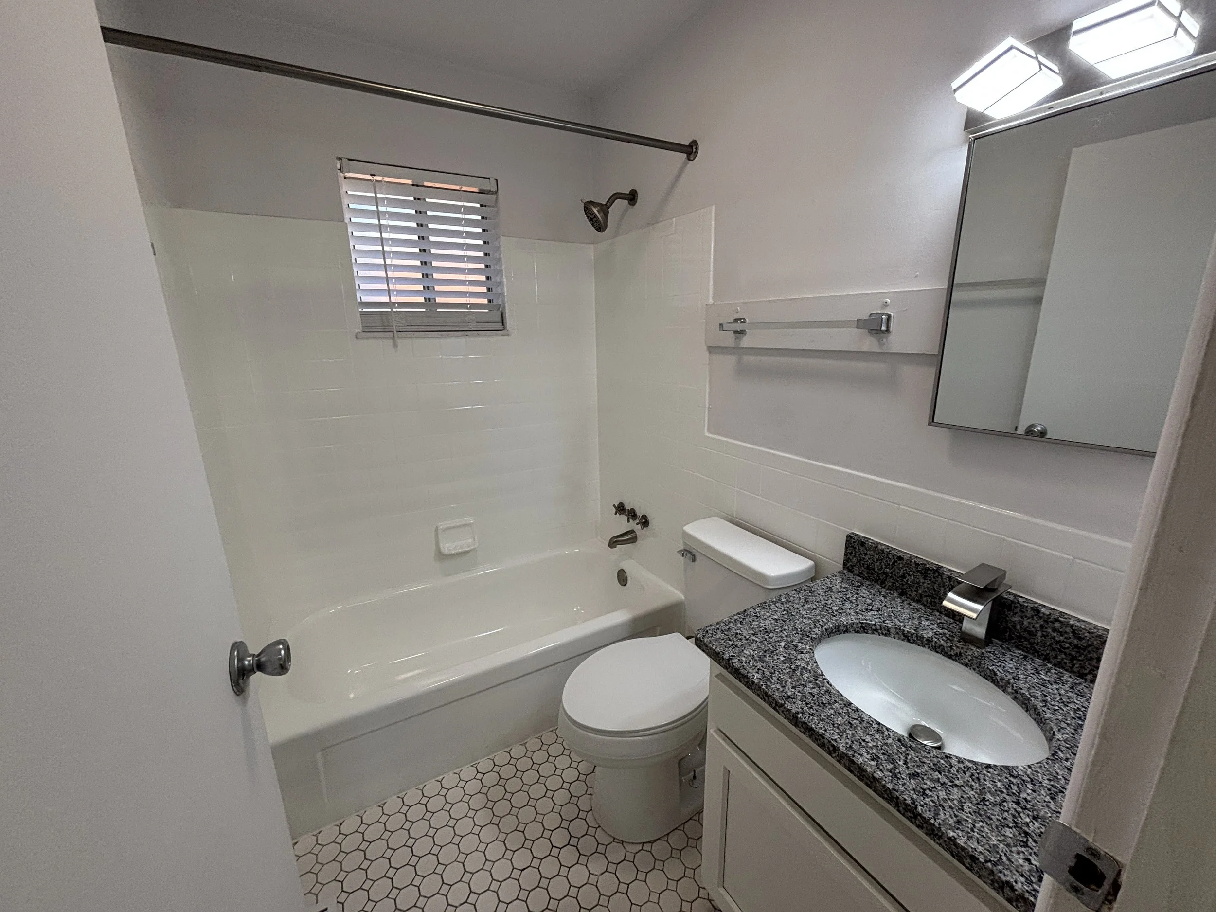 Small bathroom with white tile bathtub, toilet, granite countertop sink, mirror, small window, and light fixture.