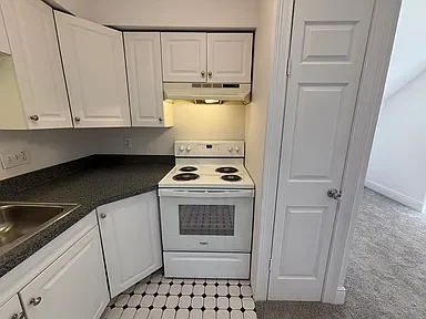 A small kitchen with white cabinets, a white stove, and a black countertop, with a partially open door leading to a carpeted room.