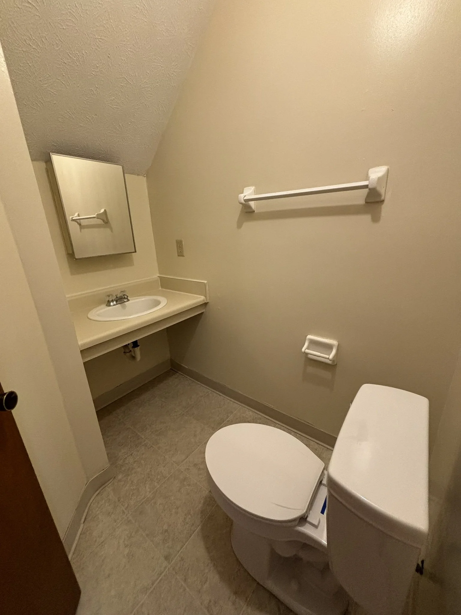 A small bathroom with a beige wall, a white toilet, a beige sink with a mirror above it, a white towel rack, and a beige toilet paper holder, with beige tiled flooring.
