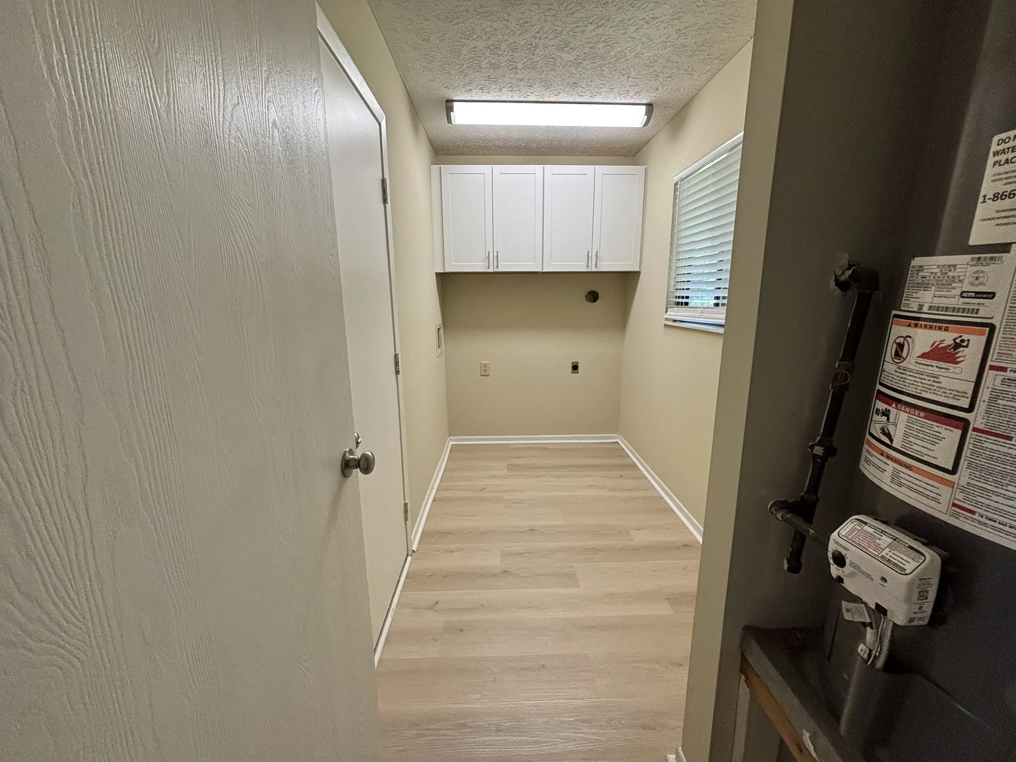 Empty laundry room with white cabinets, a window with blinds, light wood flooring, and a fluorescent ceiling light.