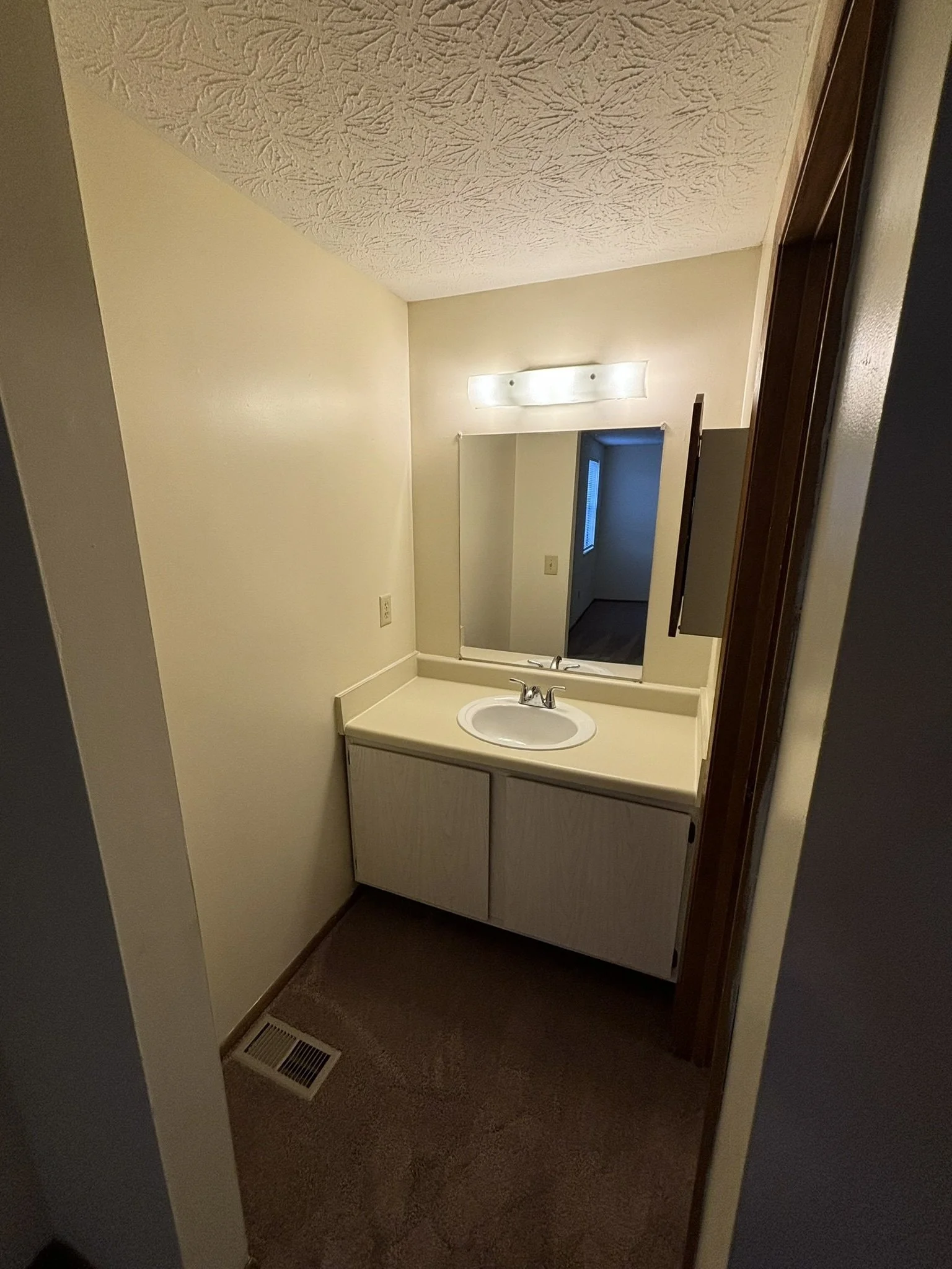 A small bathroom vanity with a white countertop, sink, and mirror. There's a wall-mounted light fixture above the mirror, beige walls, carpeted floor, and vent on the floor. A doorway in the background leads to another room with a window.