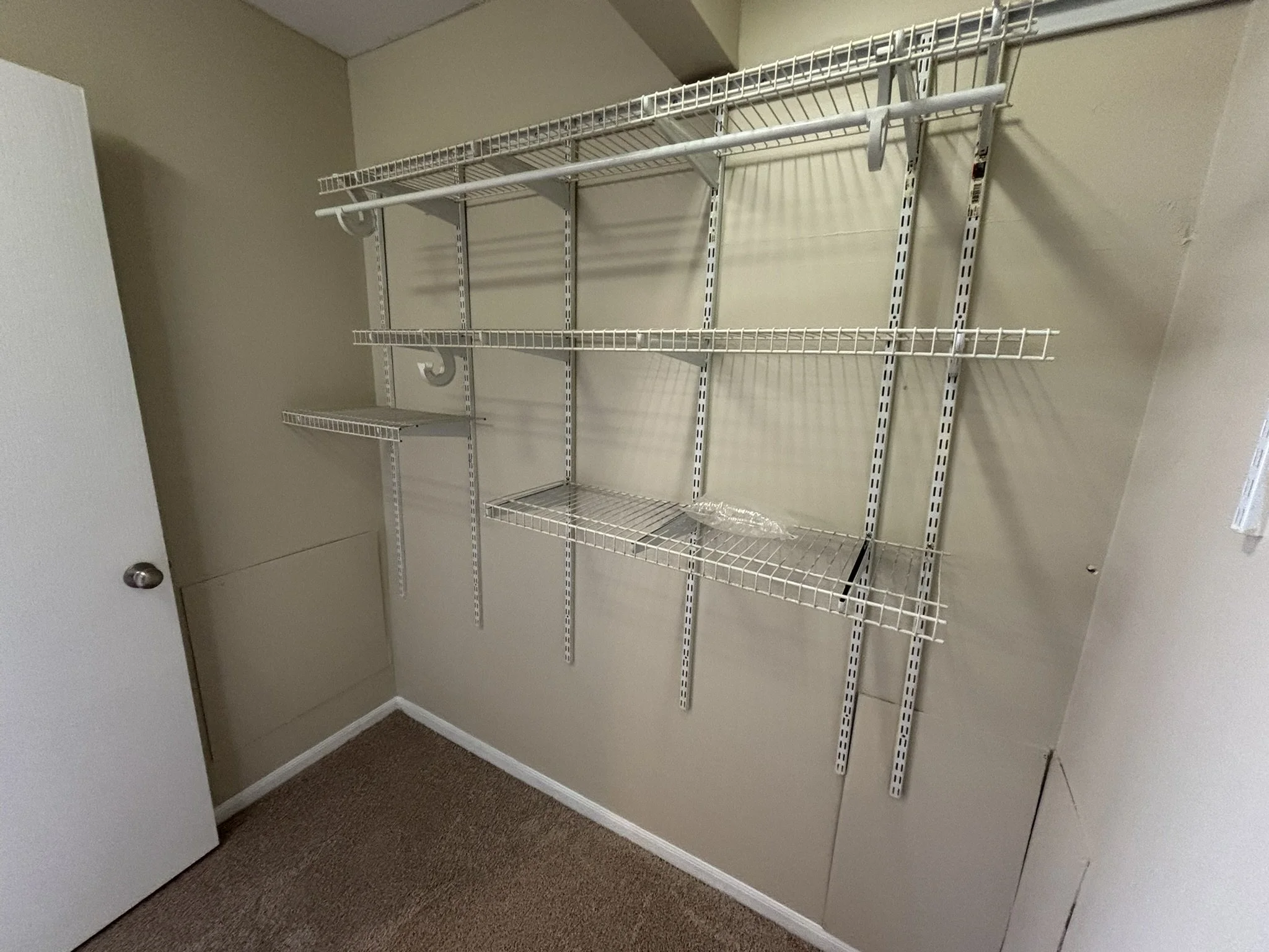 Empty white wire shelving unit attached to beige wall in a closet, with beige carpeted floor.