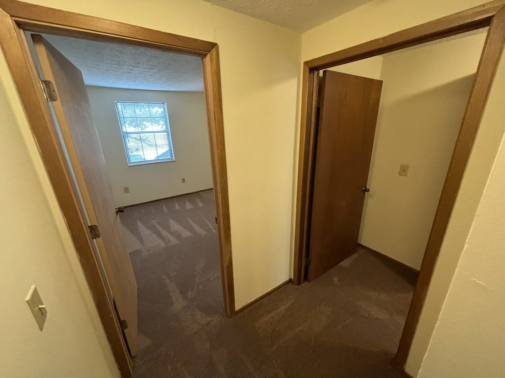 View of two rooms connected by doorways in a residential interior. Both rooms have carpeted floors, and one room has a window with blinds.