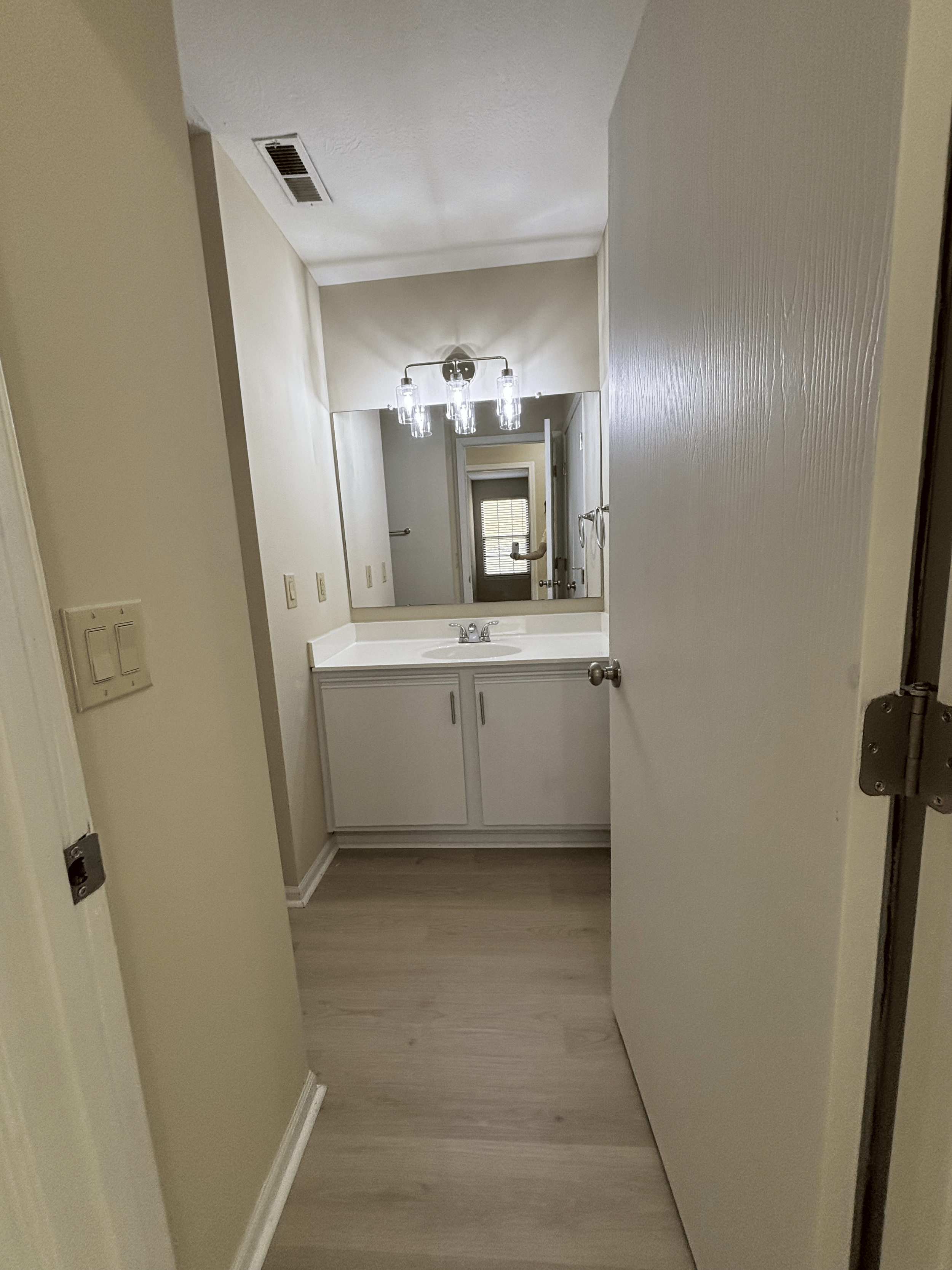 A small bathroom with a white sink vanity, a large mirror, a ceiling light fixture with multiple bulbs, and a doorway leading to a room with a window with closed blinds.