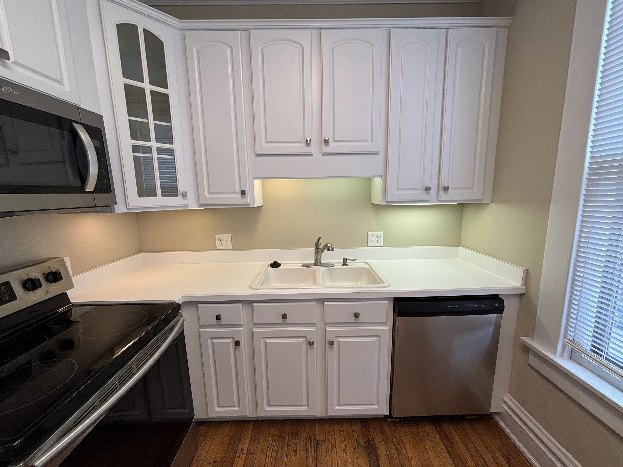 A kitchen with white cabinets, a white countertop, a stainless steel microwave, a black stovetop, a double sink, a dishwasher, and a window with blinds.