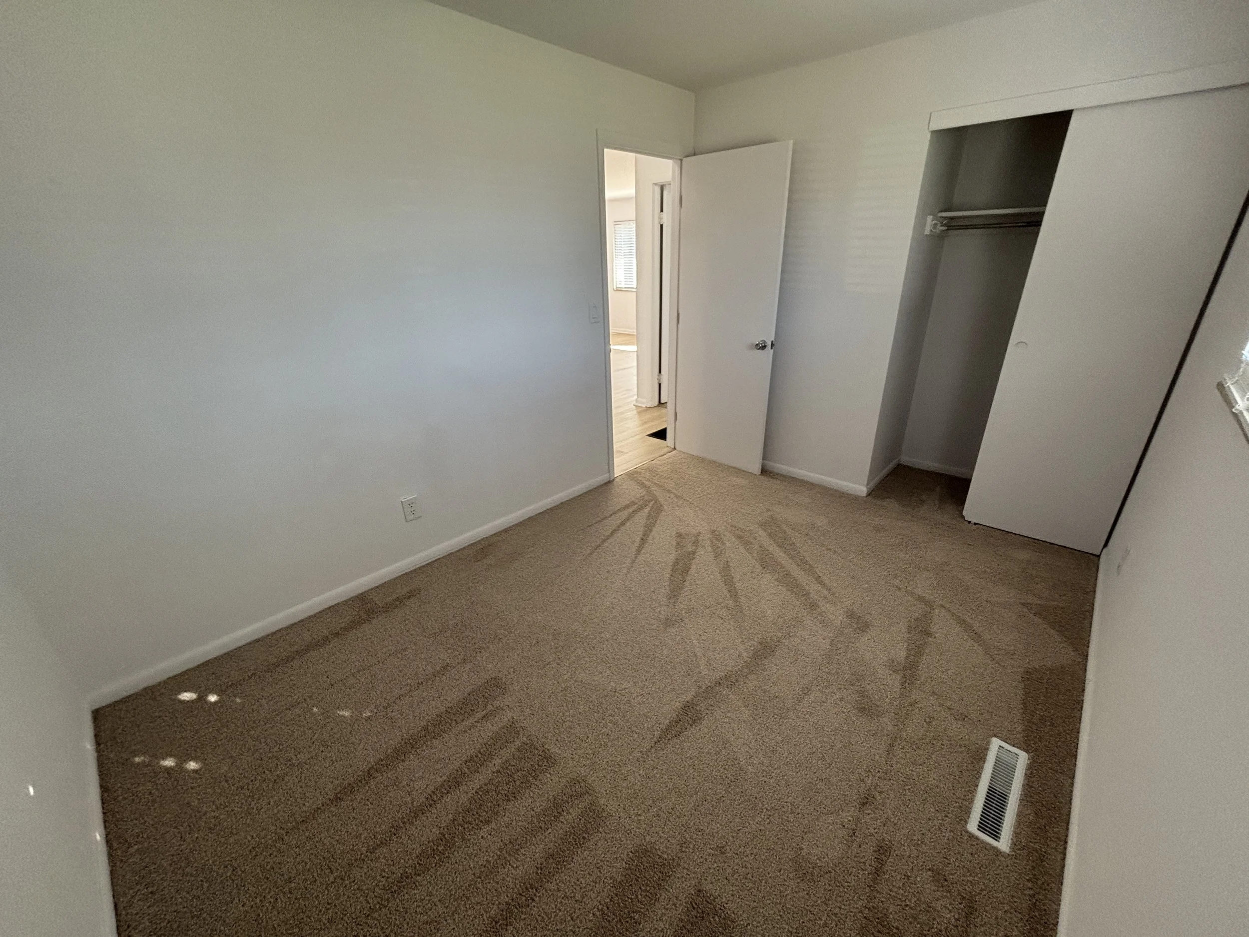 Empty bedroom with beige carpet, an open closet with sliding doors, a window, and a doorway leading to another room.