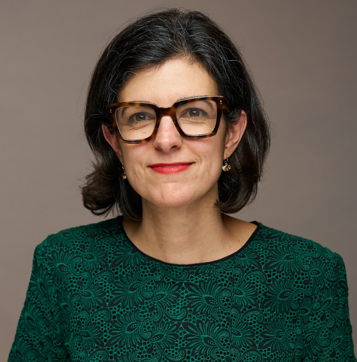 A woman with dark hair, glasses, and earrings wearing a green lace-patterned top, smiling against a neutral background.