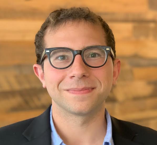 Portrait of a young man with glasses, smiling, wearing a suit jacket, with a wooden background.