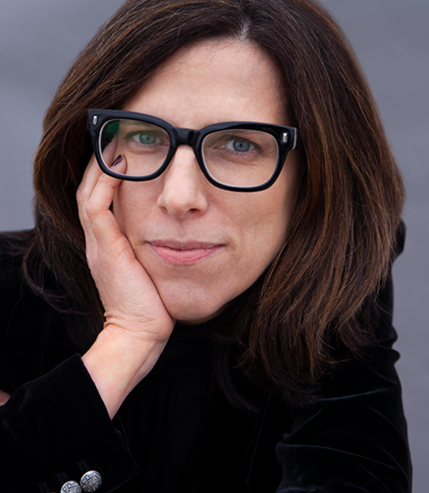 Close-up of a woman with brown hair and glasses, resting her face on her hand, looking at the camera.