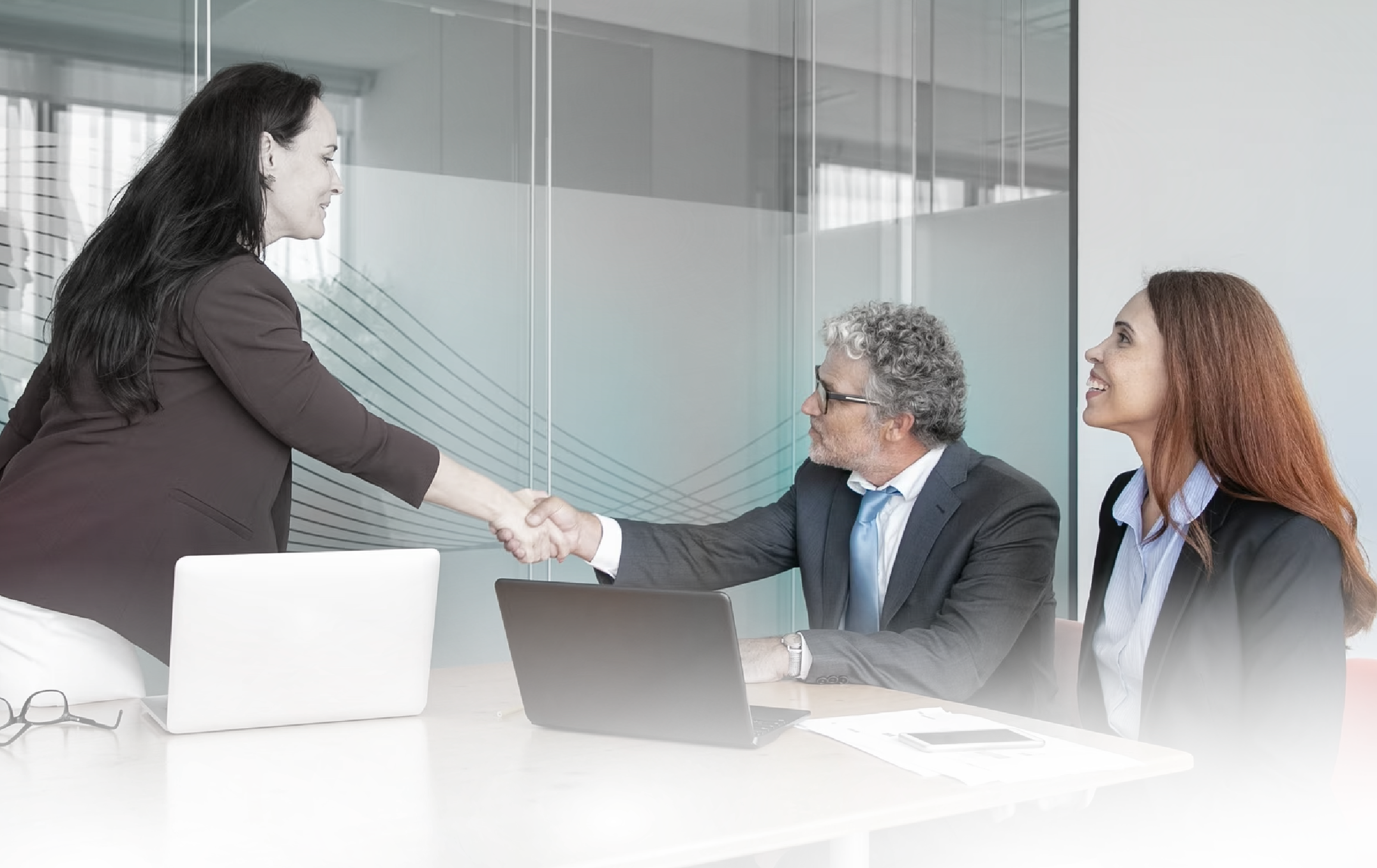 A woman in a brown blazer shaking hands with a man in glasses and a suit while another woman looks on in a modern office conference room.