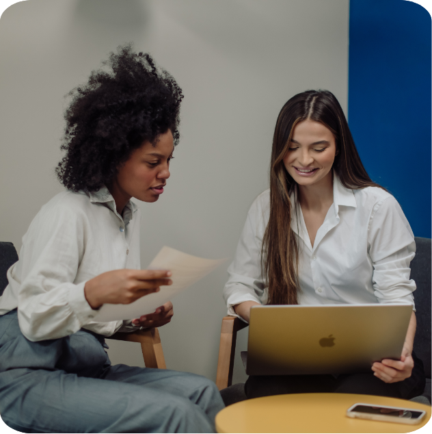 Two women sitting at a table, one holding a piece of paper and the other using a silver MacBook, engaged in a discussion in a modern office setting.