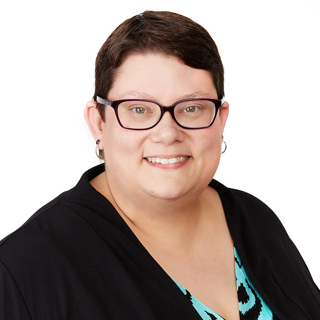 Headshot of a woman with short dark hair, wearing glasses, earrings, a black jacket, and a patterned top, smiling against a white background.