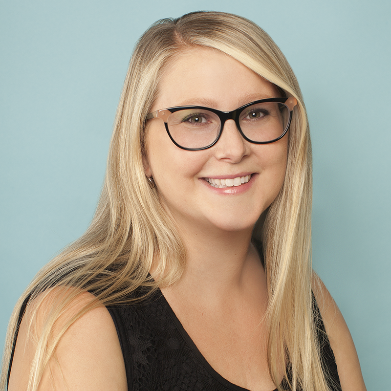 A young woman with long blonde hair, wearing glasses and a black sleeveless top, smiling against a light blue background.