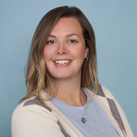 Portrait of a woman with long, wavy blond hair, smiling, wearing a light blue top and a white cardigan with gray and beige stripes, set against a blue background.