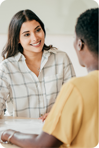 A woman with long dark hair and a checkered shirt smiling and talking to a person with short curly hair and a yellow shirt, in a professional or counseling setting.