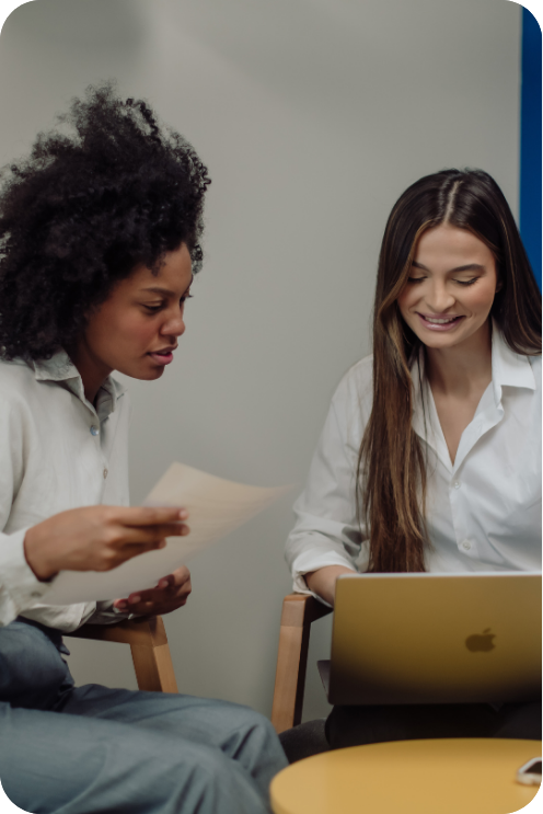 Two women sitting at a table, one with curly black hair looking at a paper, the other with long brown hair using a laptop, in a casual office setting.
