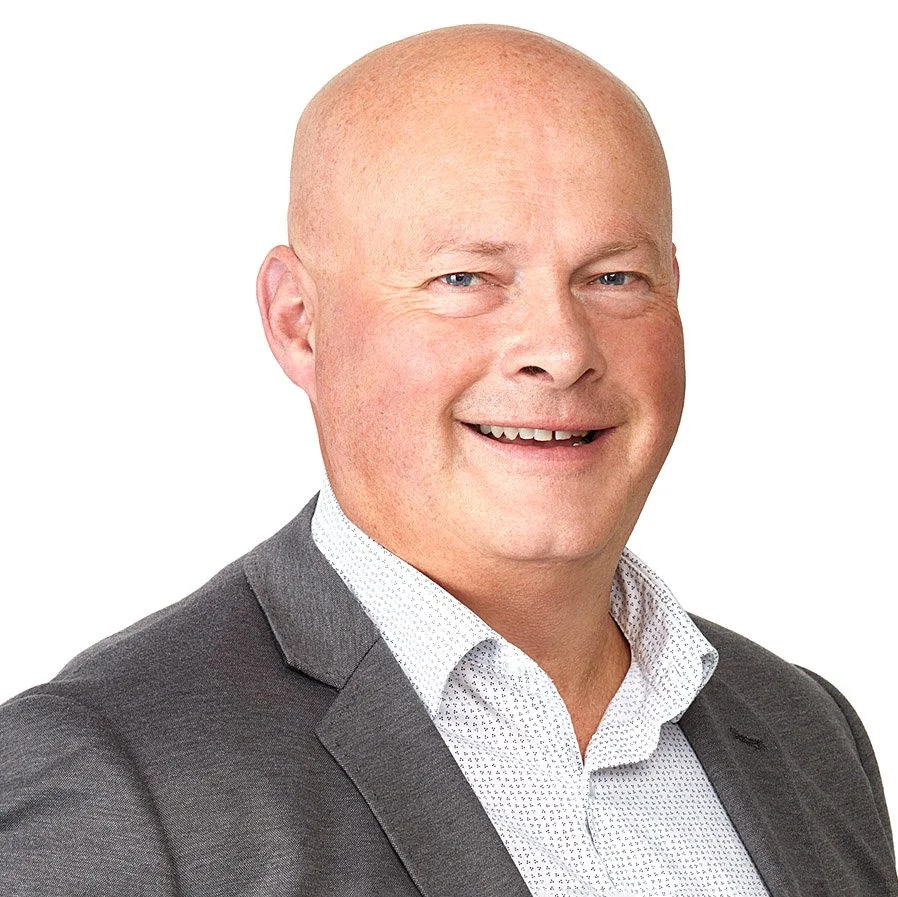 A bald man in a gray suit with a white patterned shirt smiling against a white background.