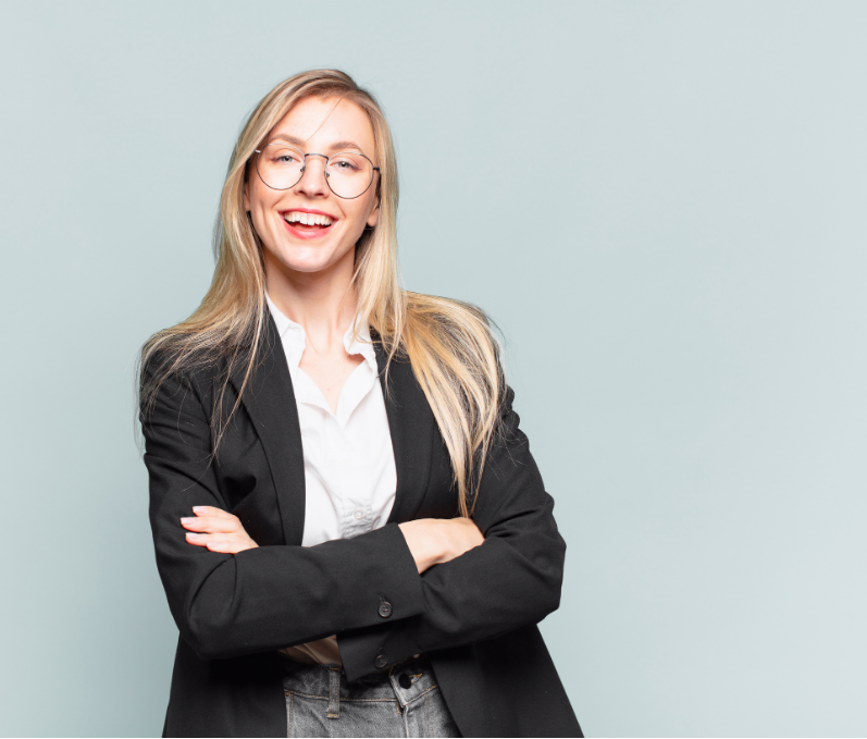A smiling woman with blonde hair, wearing glasses, a black blazer, and a white shirt, standing with her arms crossed against a light blue background.