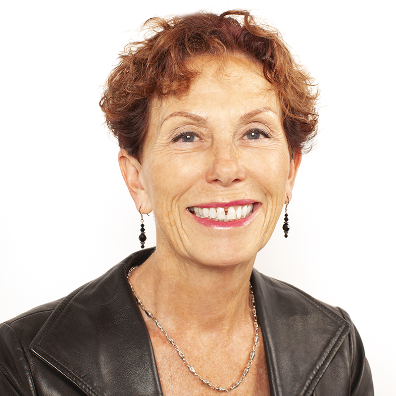 Close-up portrait of a cheerful middle-aged woman with short curly auburn hair, wearing earrings, a necklace, and a black leather jacket against a white background.