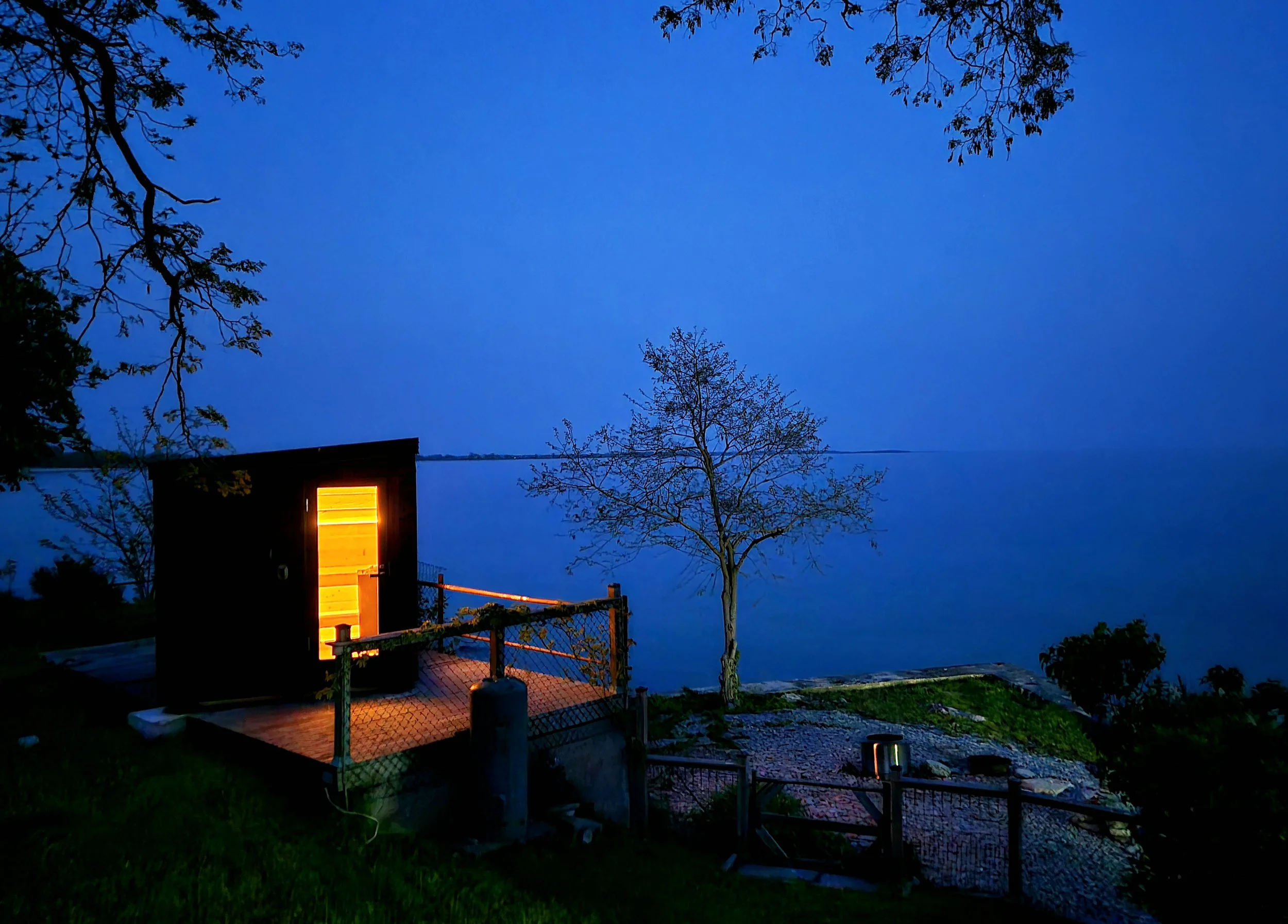 A small illuminated cabin on a lakeside at dusk with a single tree and a wooden fence in the foreground.