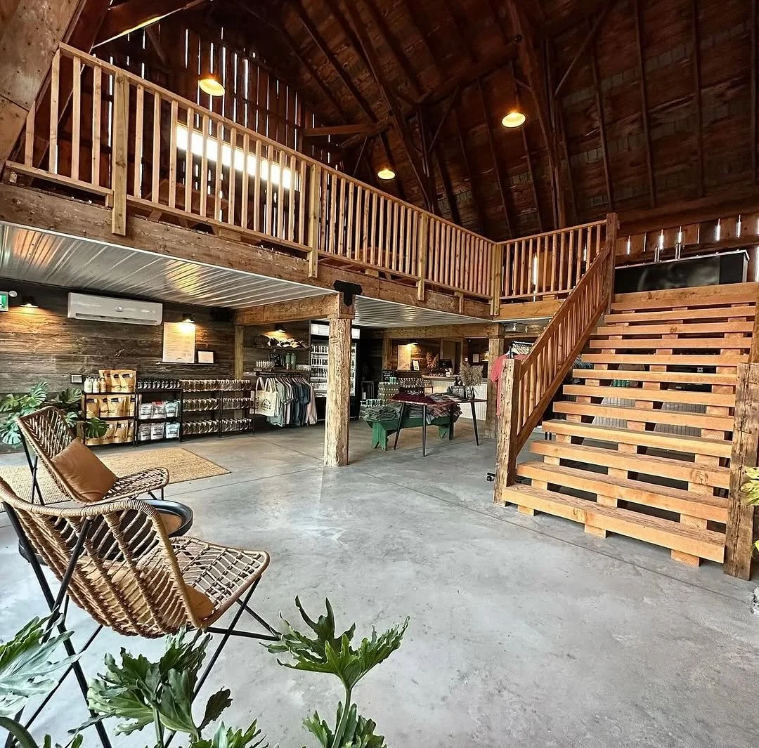 Interior of a rustic shop or cafe with wooden accents, high vaulted ceiling, and a mezzanine level accessible via wooden stairs. There are wicker chairs with cushions, retail shelves with products, hanging lights, and plants.