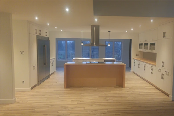 Modern, empty kitchen with wooden floors, white cabinets, a large island, and a stainless steel range hood, with large windows at the back.