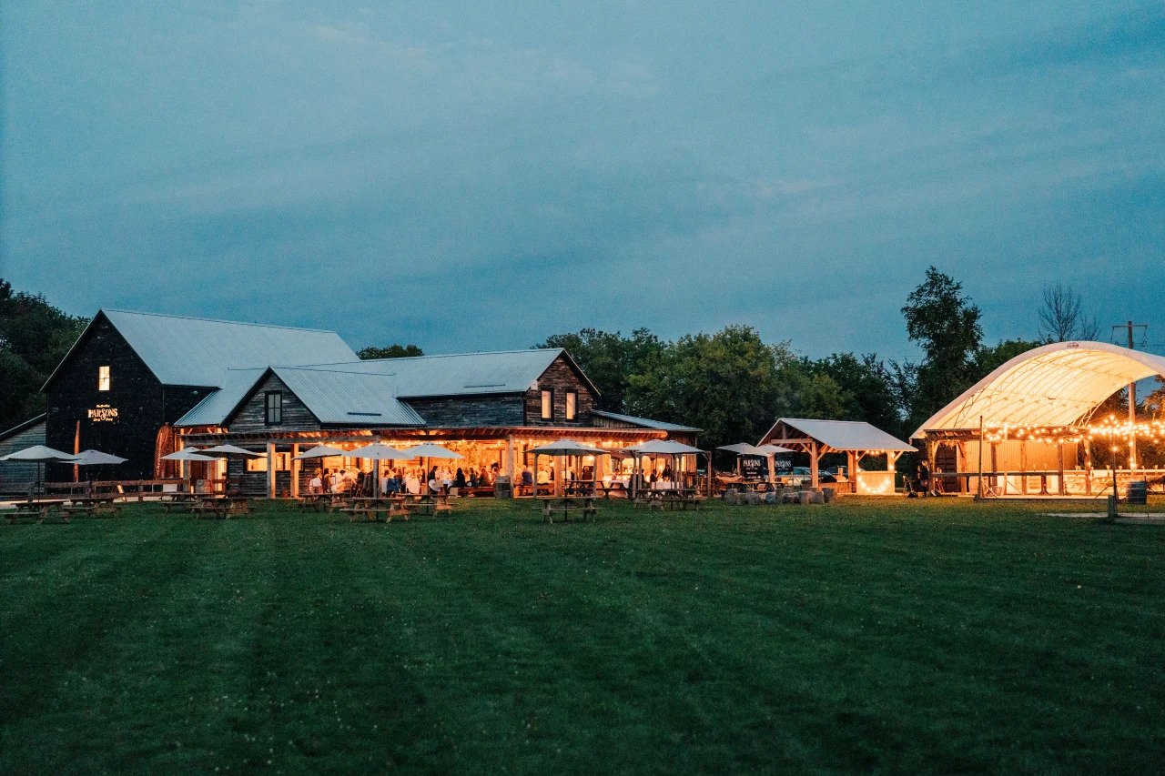 A rustic outdoor event space with a barn and a pavilion illuminated with string lights, surrounded by green lawn and trees at dusk.