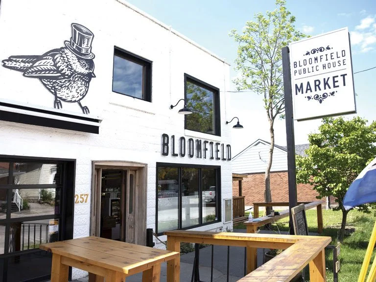 Exterior of Bloomfield Public House, a building with a mural of a bird wearing a top hat, the word 'BLOOMFIELD' on the wall, and outdoor seating with wooden tables. A sign indicates it is a market.