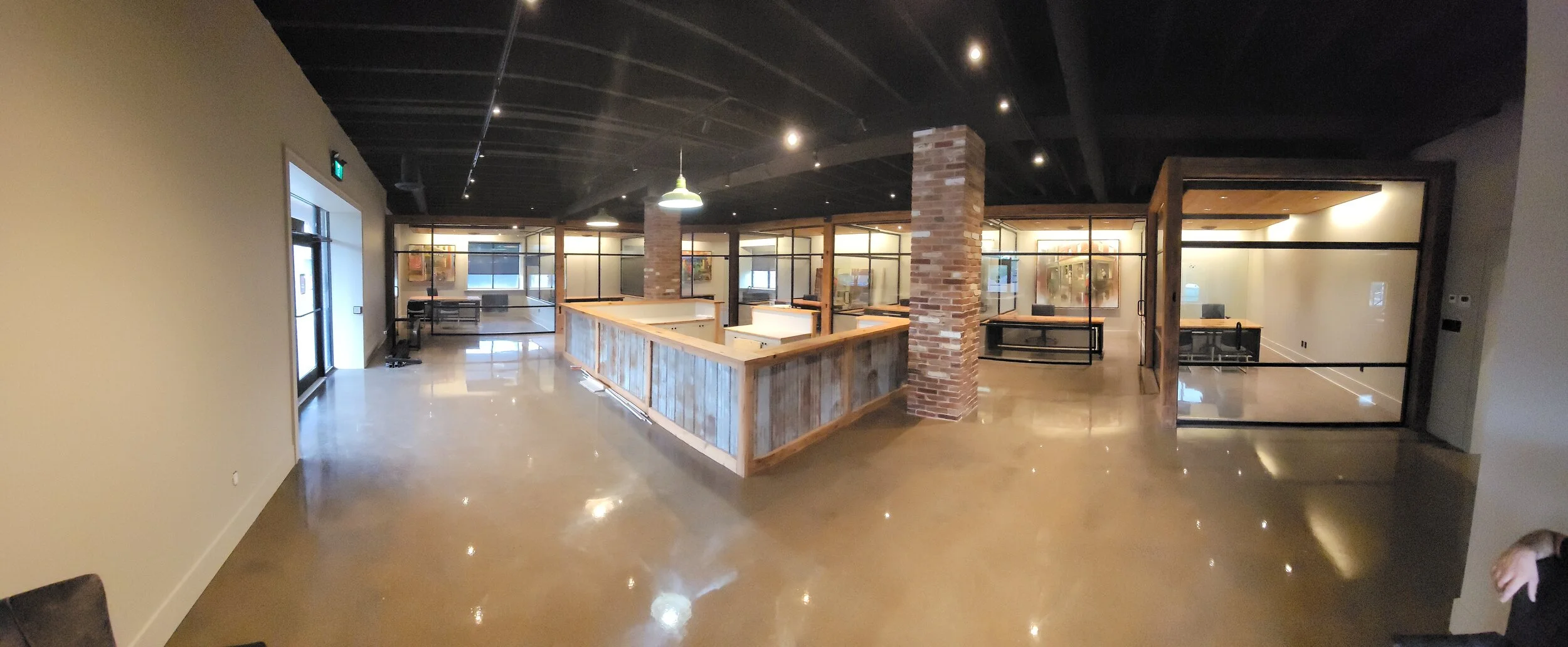 Interior of an office space with an open layout, featuring desks with computers, glass partition walls, exposed brick pillars, and a polished concrete floor.