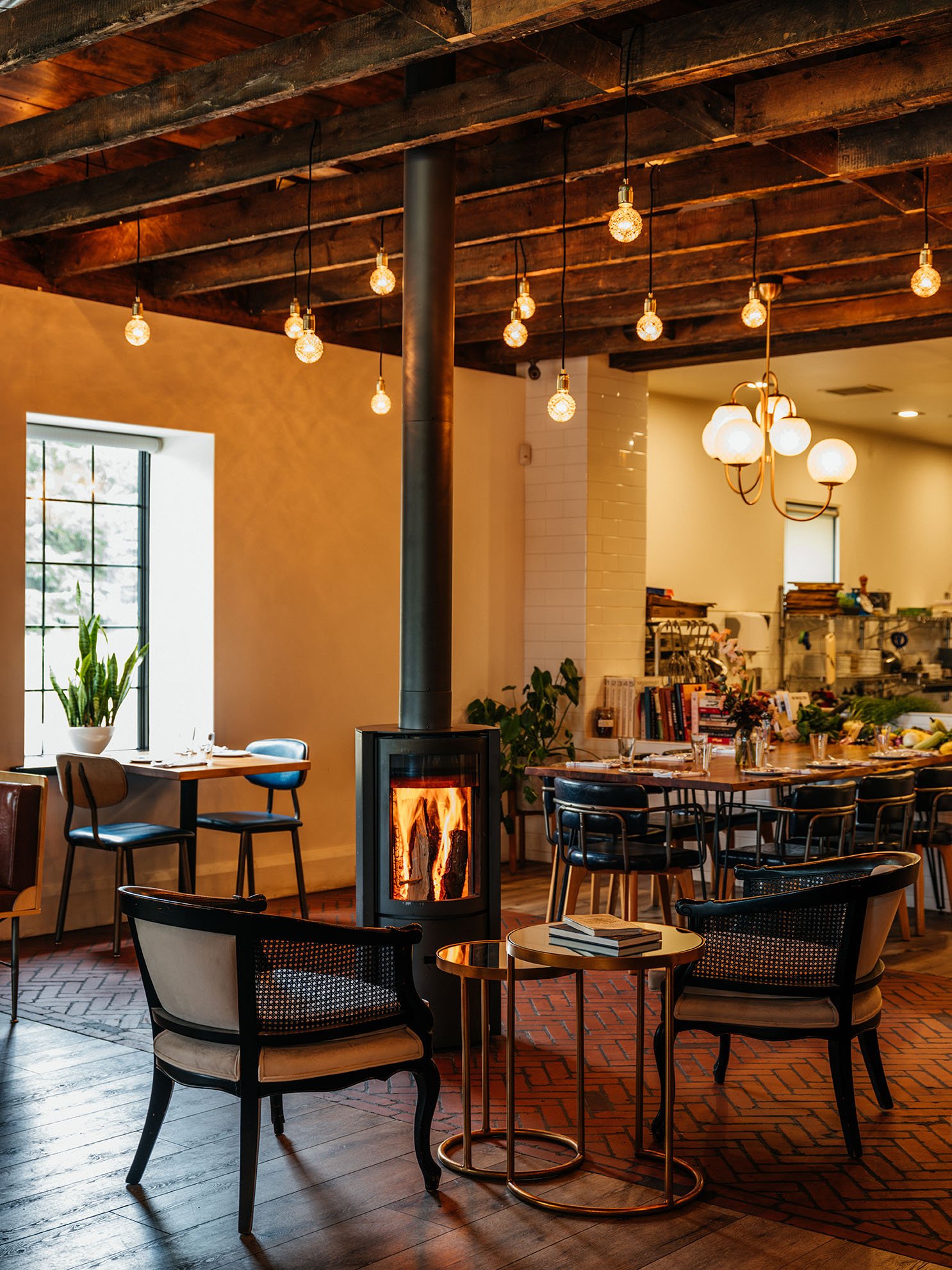 Cozy dining area with a wood-burning stove, string lights, and various tables and chairs in a warmly lit restaurant.