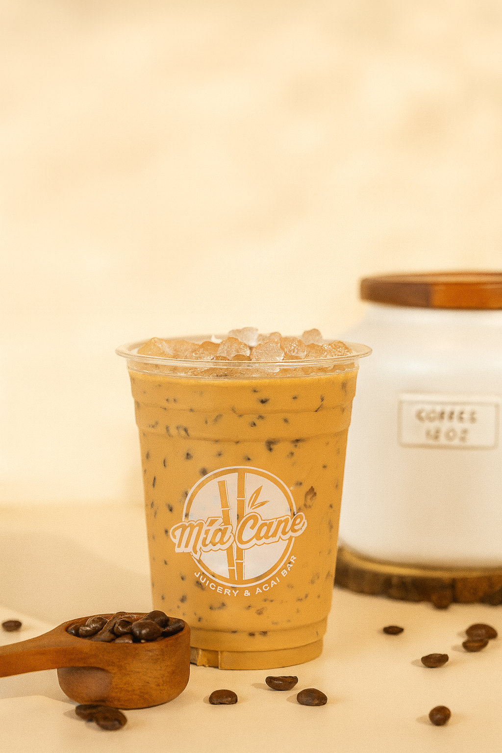 An iced coffee beverage in a clear cup with a logo reading 'Mia Cane Juicery & Acai Bar,' topped with ice and surrounded by scattered coffee beans and a small wooden scoop filled with coffee beans. A white jar with a wooden lid labeled 'COFFEE HOT' i