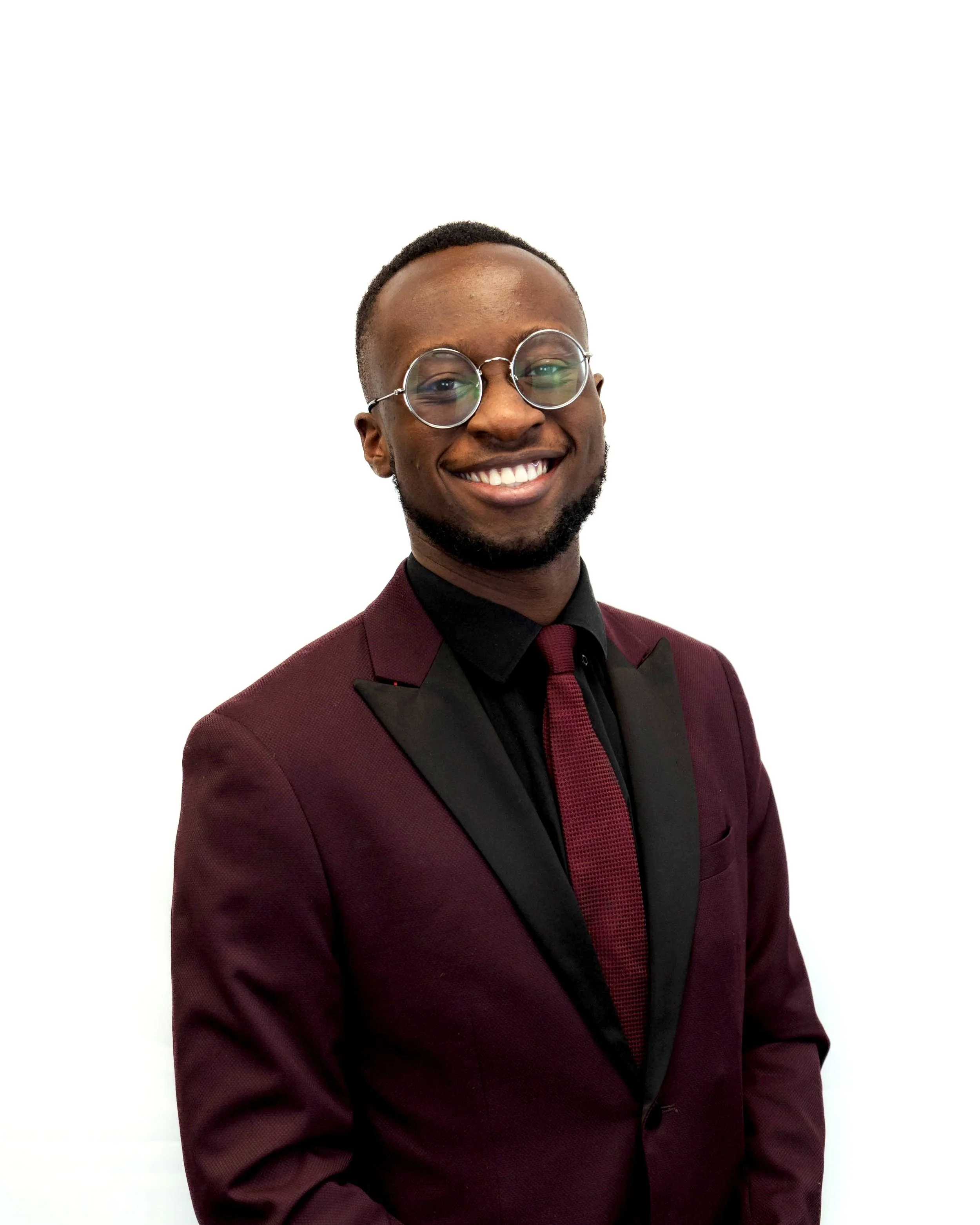 Smiling young man in a maroon suit, black shirt, maroon tie, and round glasses posing against a white background.