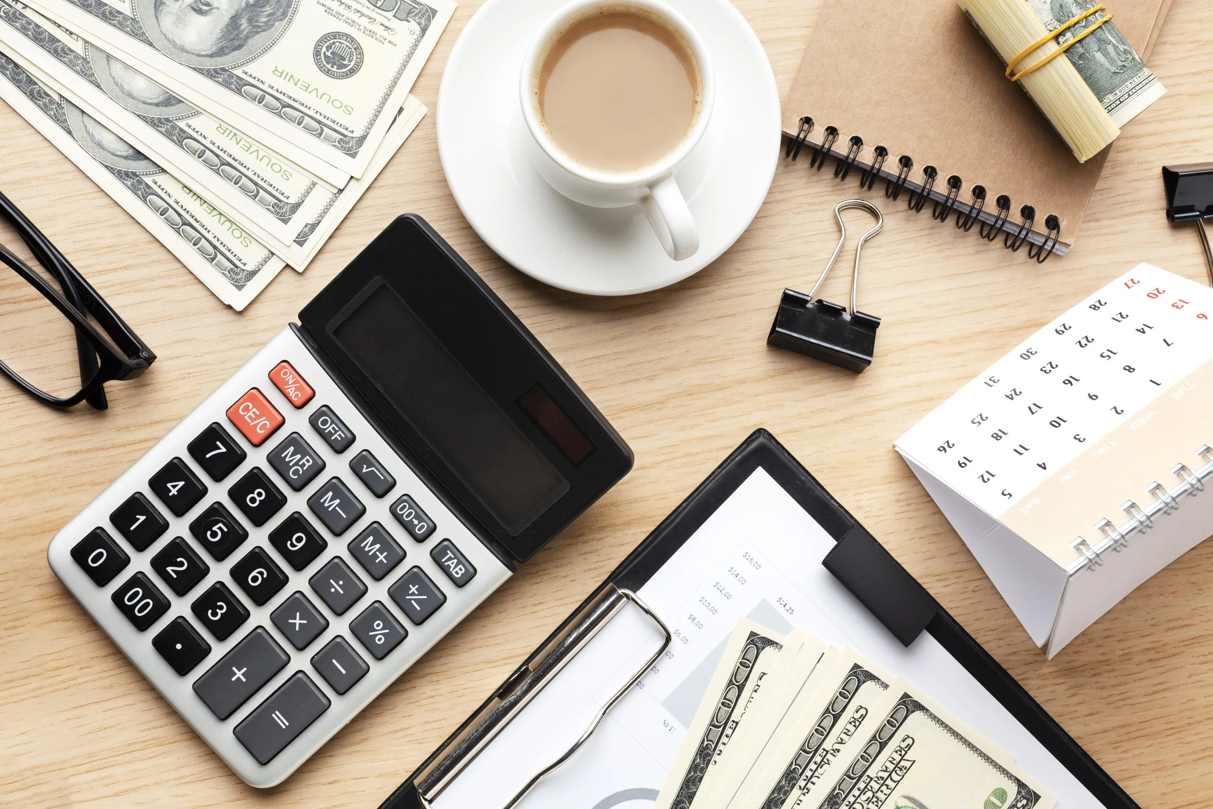 Office desk with a calculator, a cup of coffee, rolled dollar bills, a notepad, a clipboard with a receipt and cash, a pair of glasses, and a calendar.
