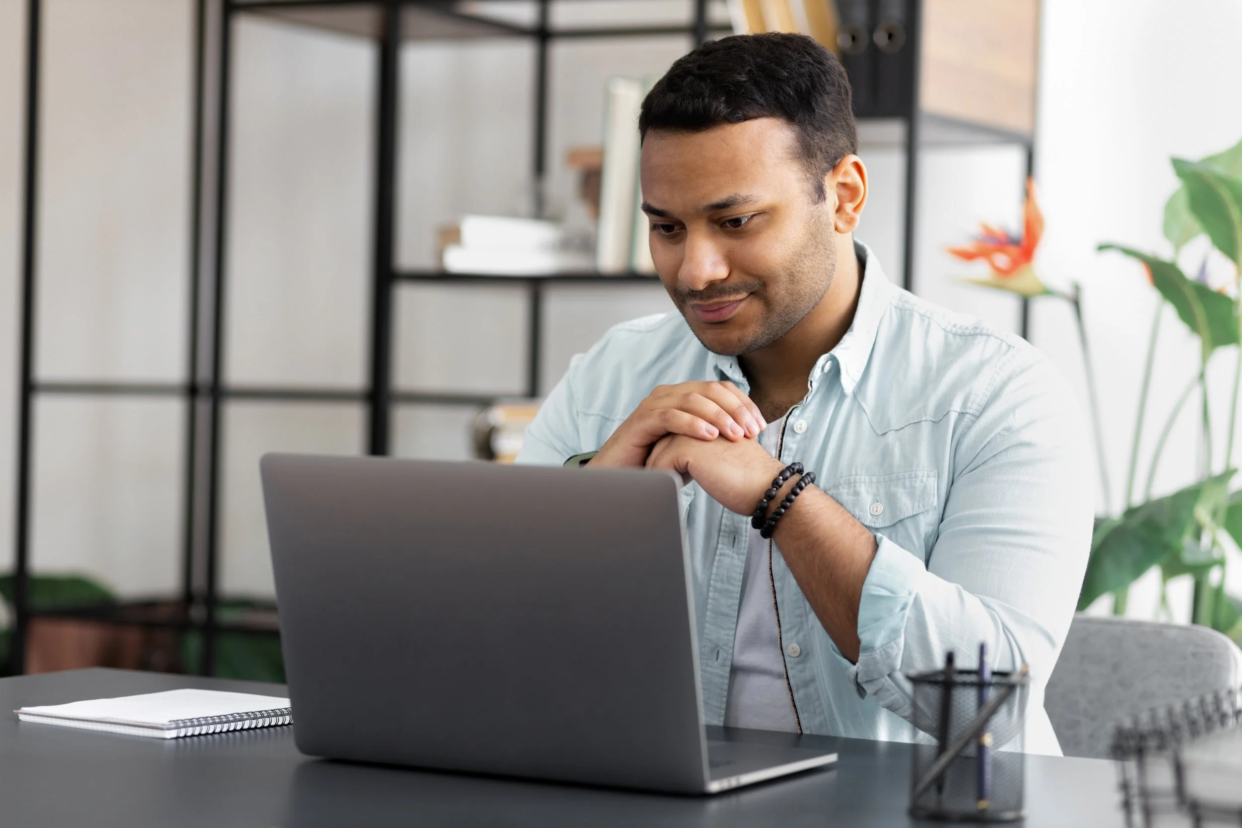 A young man sitting at a desk with a laptop, looking down at the screen with hands clasped, in a modern office or home workspace, with books and plants in the background.