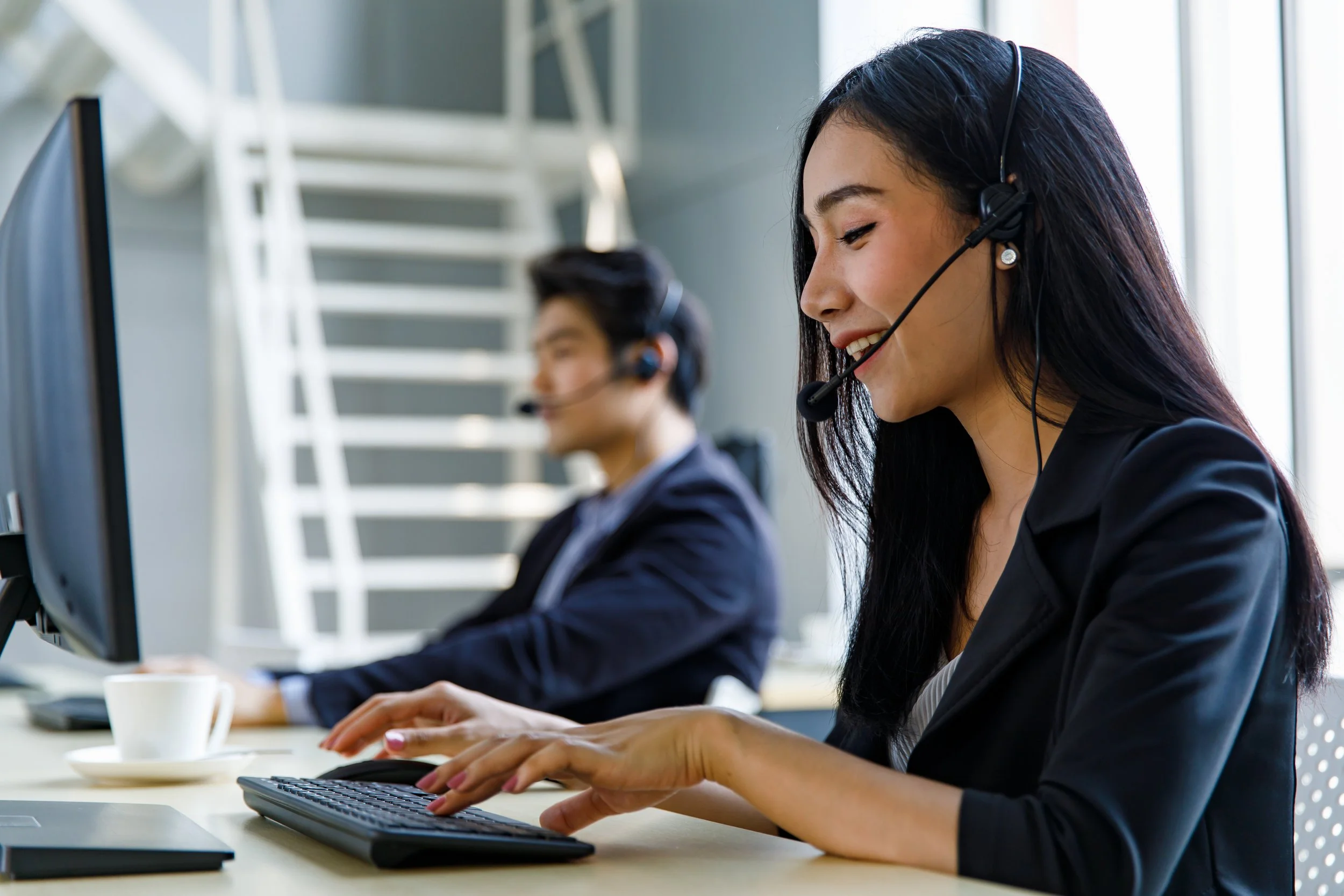 Two customer service representatives, a woman in the foreground and a man in the background, wearing headsets and working at computers in an office setting.