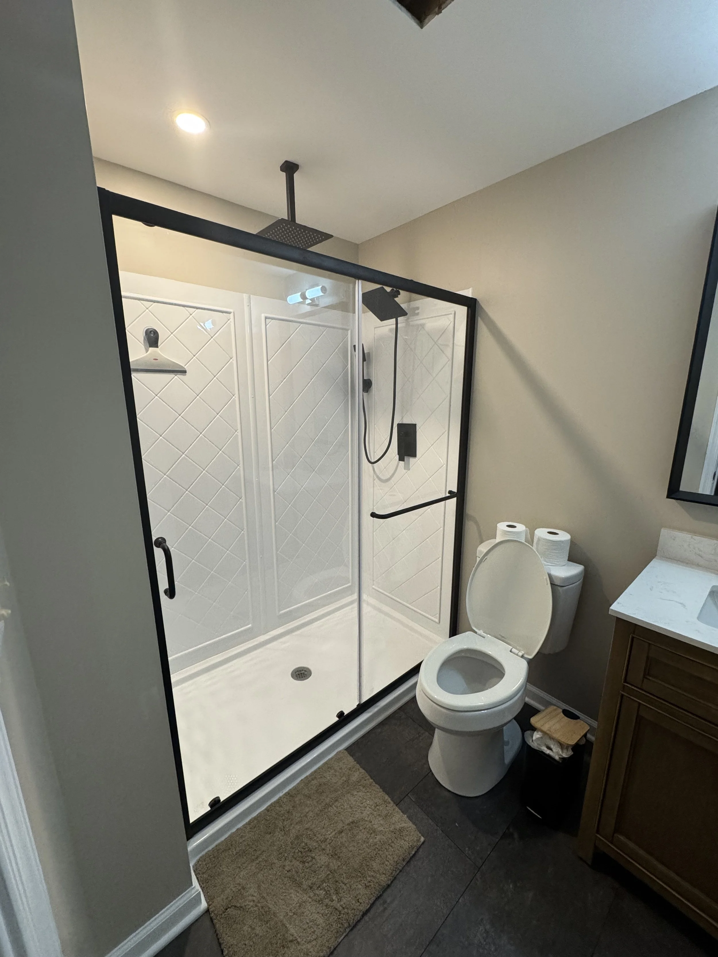 A modern bathroom with a glass-enclosed shower, a toilet with two rolls of toilet paper on top, a wooden cabinet with a white countertop and sink, a mirror, a small trash bin, and a beige rug on dark tile flooring.