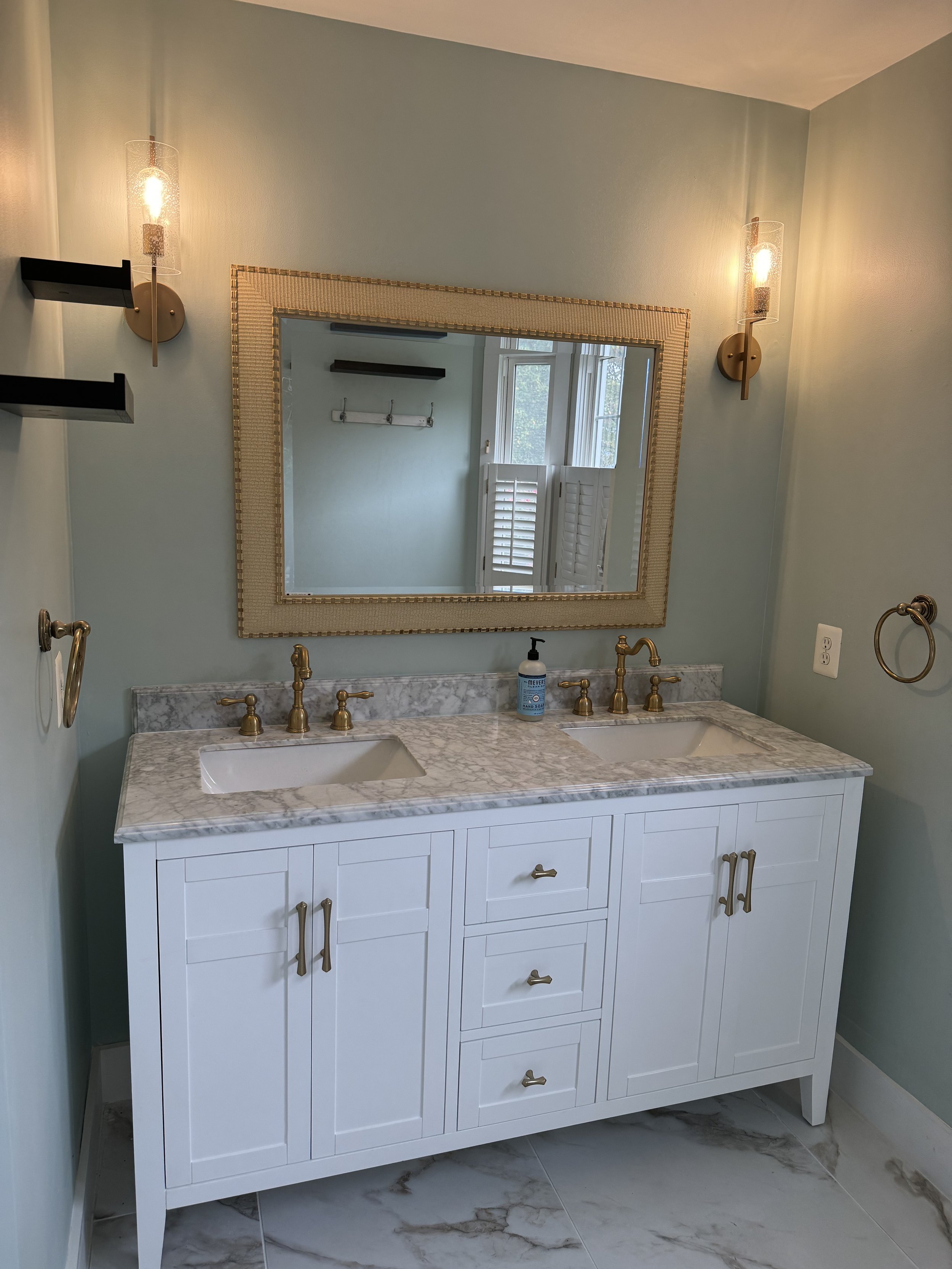 Modern bathroom vanity with twin sinks, white cabinets, and a marble countertop, complemented by gold fixtures and a large mirror.