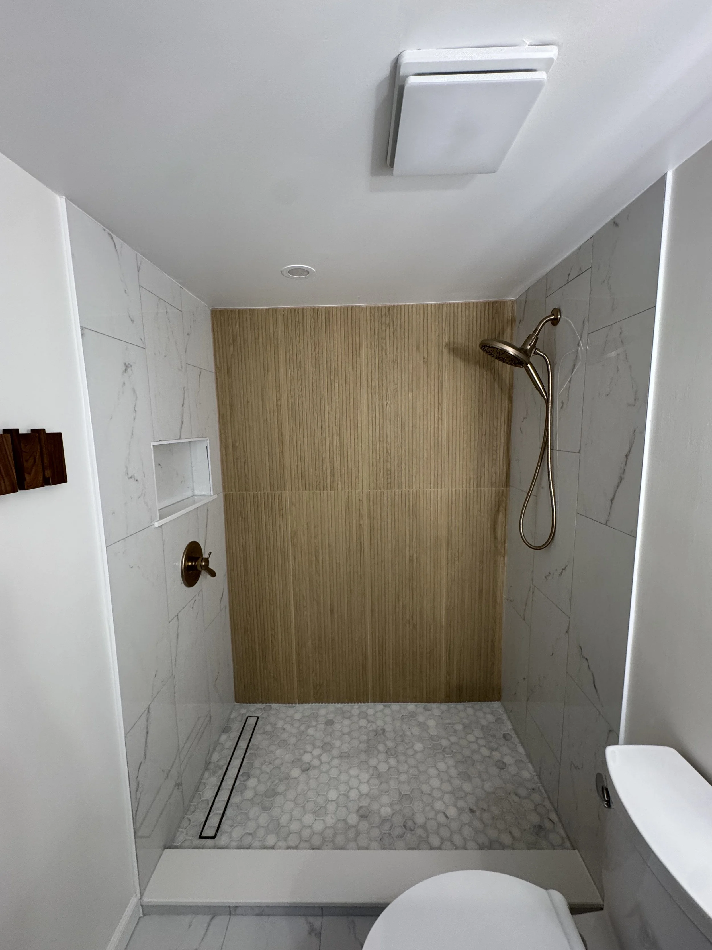 Modern walk-in shower with white marble tiles, a wooden wall panel at the back, a bronze showerhead, and a niche for toiletries. The shower area has a hexagonal tile floor and a linear drain.