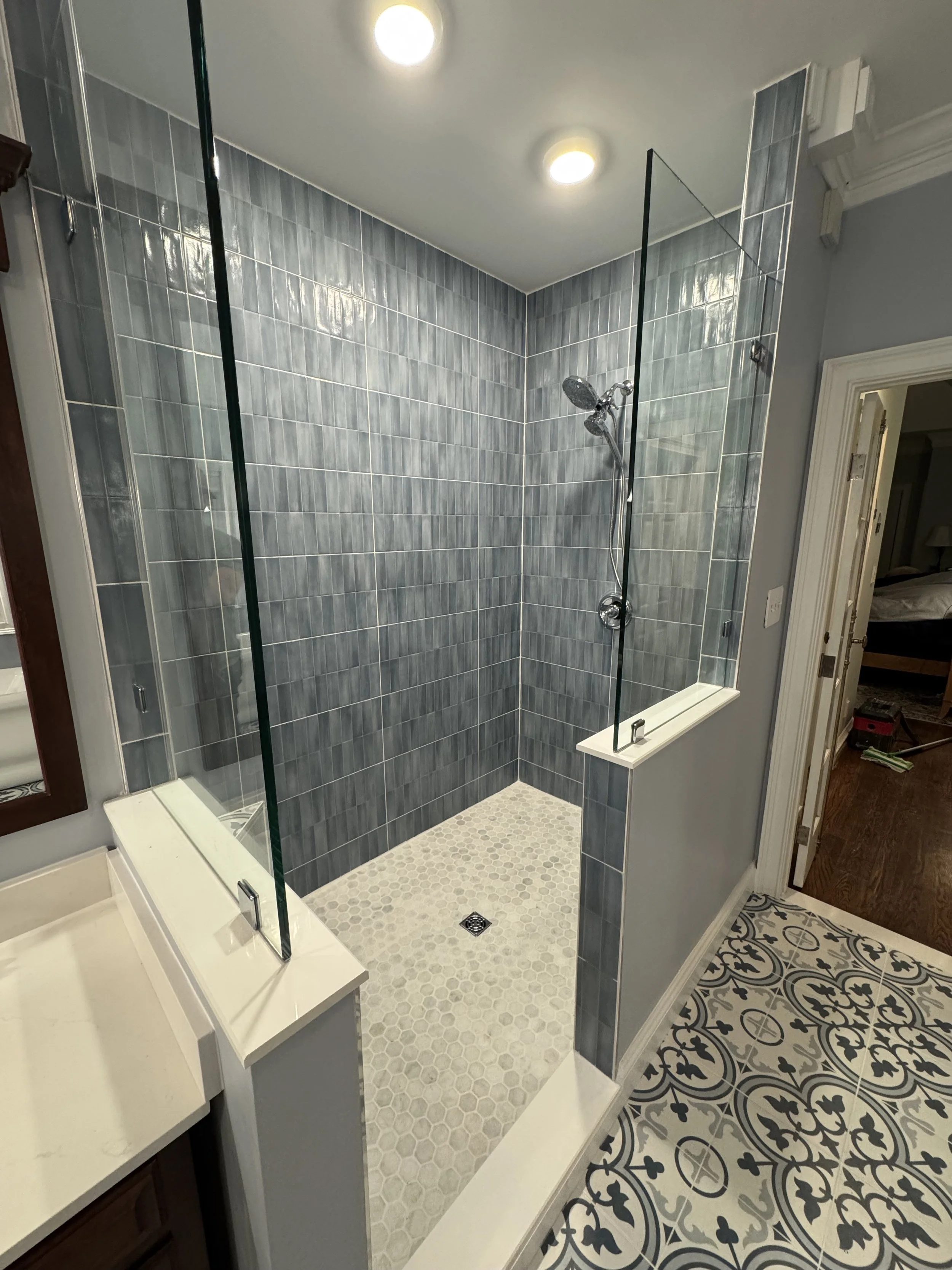 A shower area with gray tiled walls, a glass partition, and a handheld showerhead, adjacent to a patterned tile floor.