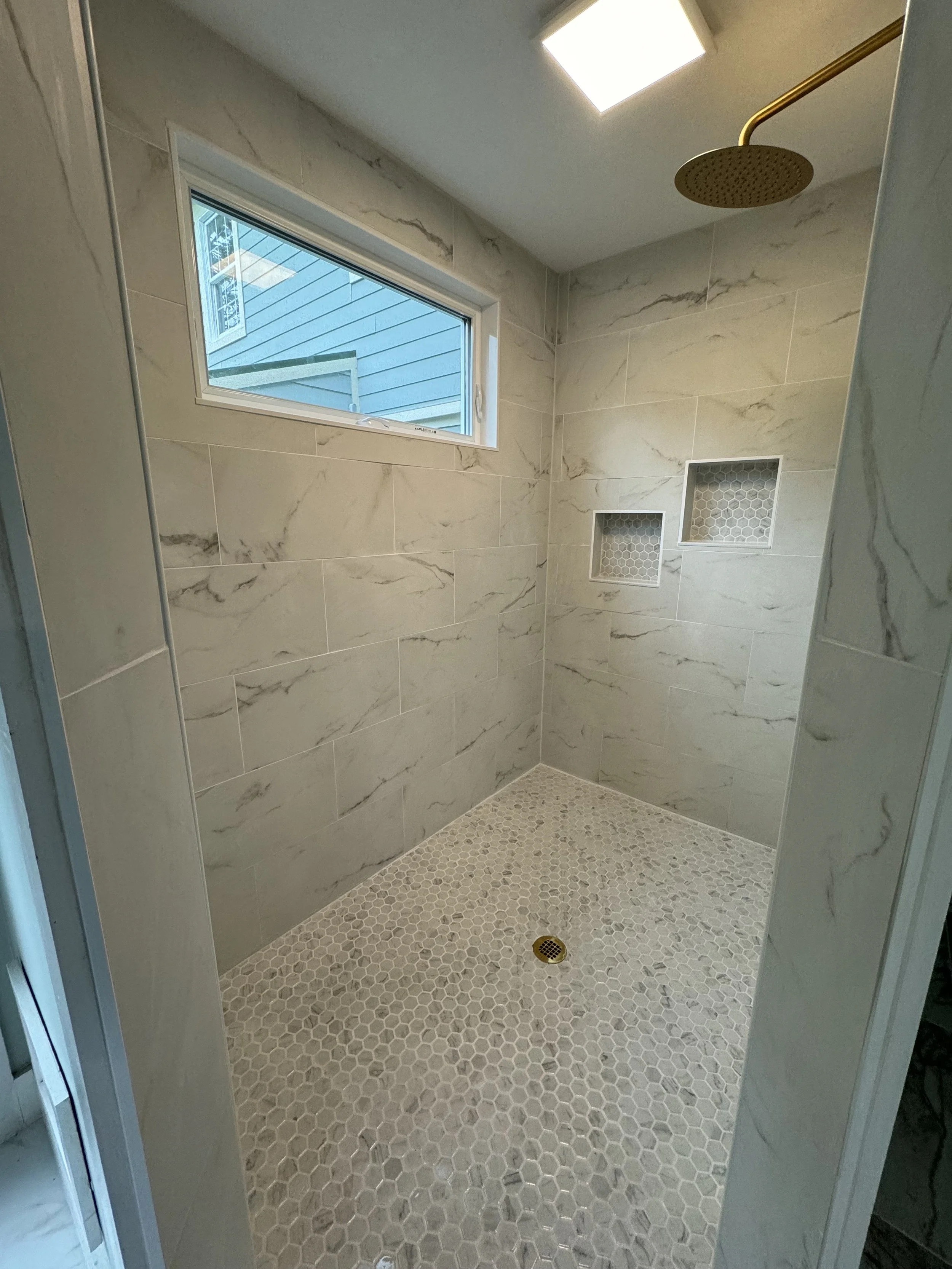 Empty walk-in shower with beige marble tiles, small hexagonal floor tiles, two built-in wall niches with hexagonal tile backs, a small horizontal window, and a ceiling-mounted rainfall showerhead.