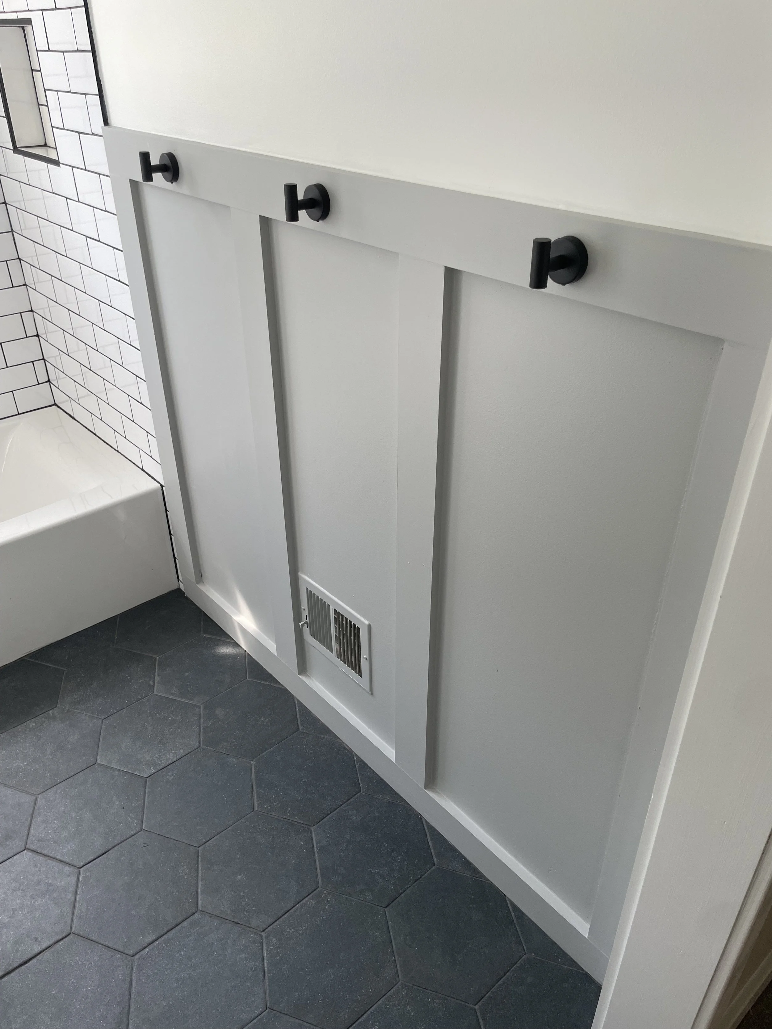 White cabinet with three black hooks on a white wall in a bathroom, with a hexagonal dark gray tiled floor and part of a bathtub with white subway tile wall.