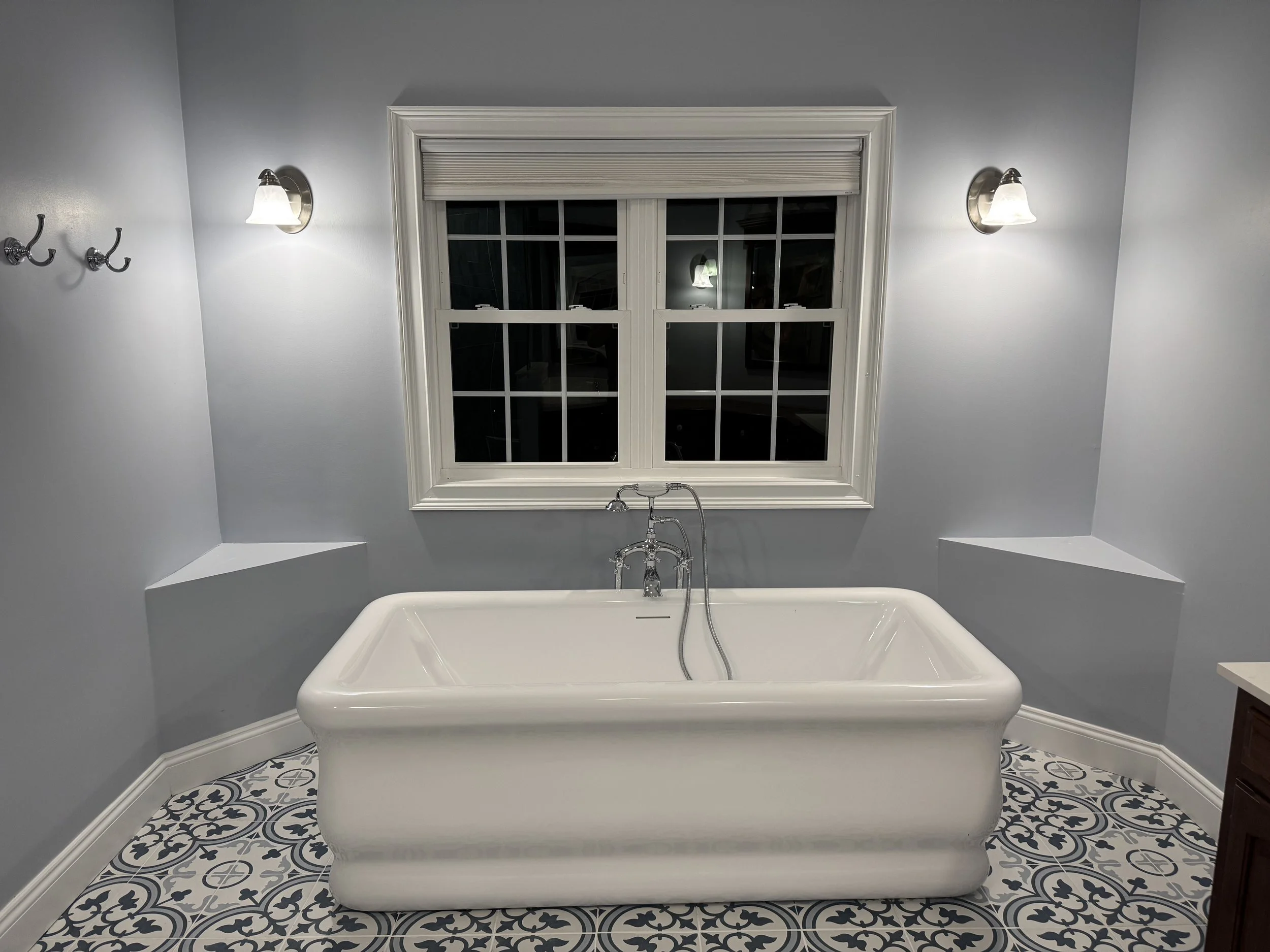 A bathroom with a white clawfoot bathtub, a large window above it, gray walls, decorative black and white tile floor, and wall-mounted light fixtures.
