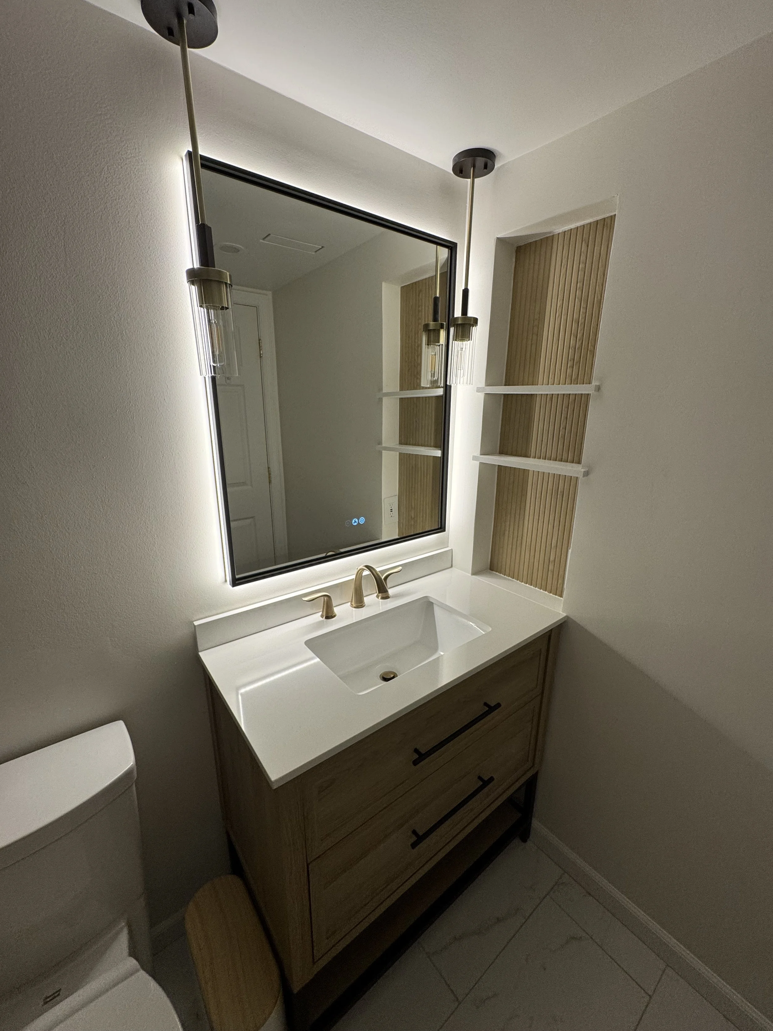 Bathroom vanity with a white countertop, a rectangular sink, and dual gold faucets, reflected in a large backlit mirror. Two hanging pendant lights with clear glass and gold accents are above the mirror. To the right of the mirror, there are three wh
