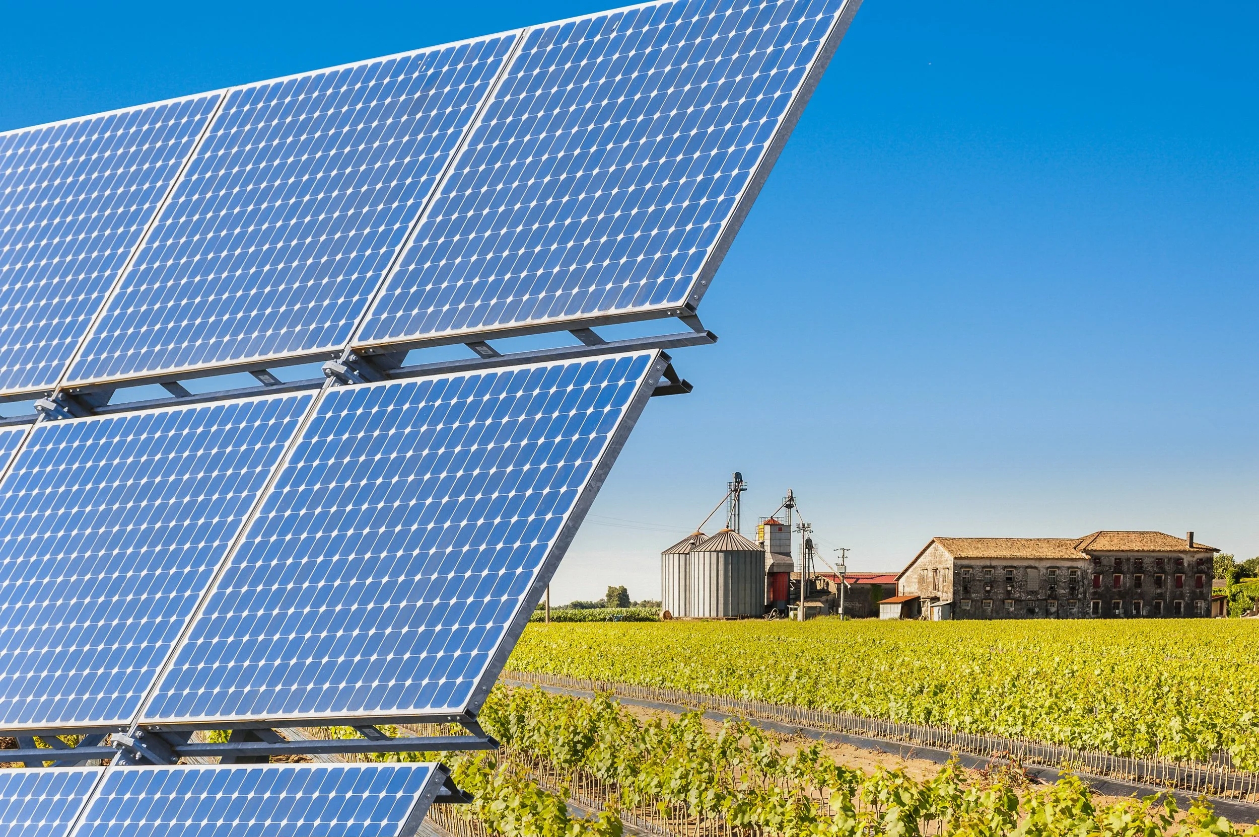Solar panels installed on a rural farm with green fields, farm buildings, and silos in the background under a clear blue sky.