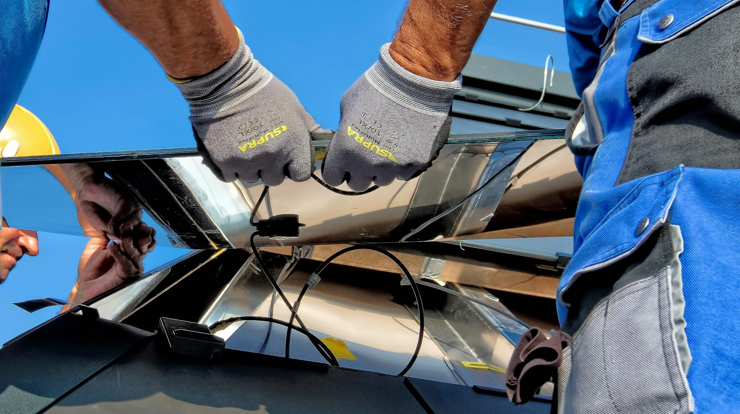 Two individuals working on a large outdoor antenna or satellite dish; one person's hands are visible wearing gray gloves with yellow text, while they work on the structure under a clear blue sky.